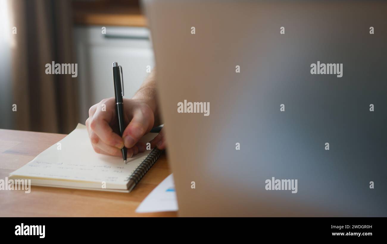 Nahaufnahme eines Mannes, der zu Hause in der Küche mit seinem Laptop arbeitet, Notizen macht und recherchiert. Viel beschäftigter Freelancer, der an modernen Tech-Notebook-devi arbeitet Stockfoto
