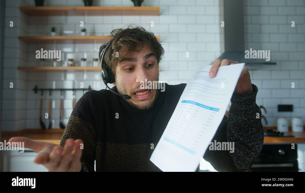 Junger Mann mit schnurlosem Headset, der zu Hause in der modernen Küche sitzt, redet, Berichte in Videokonferenzgesprächen zeigt, wütend auf das Personal, wirft Papiere Stockfoto