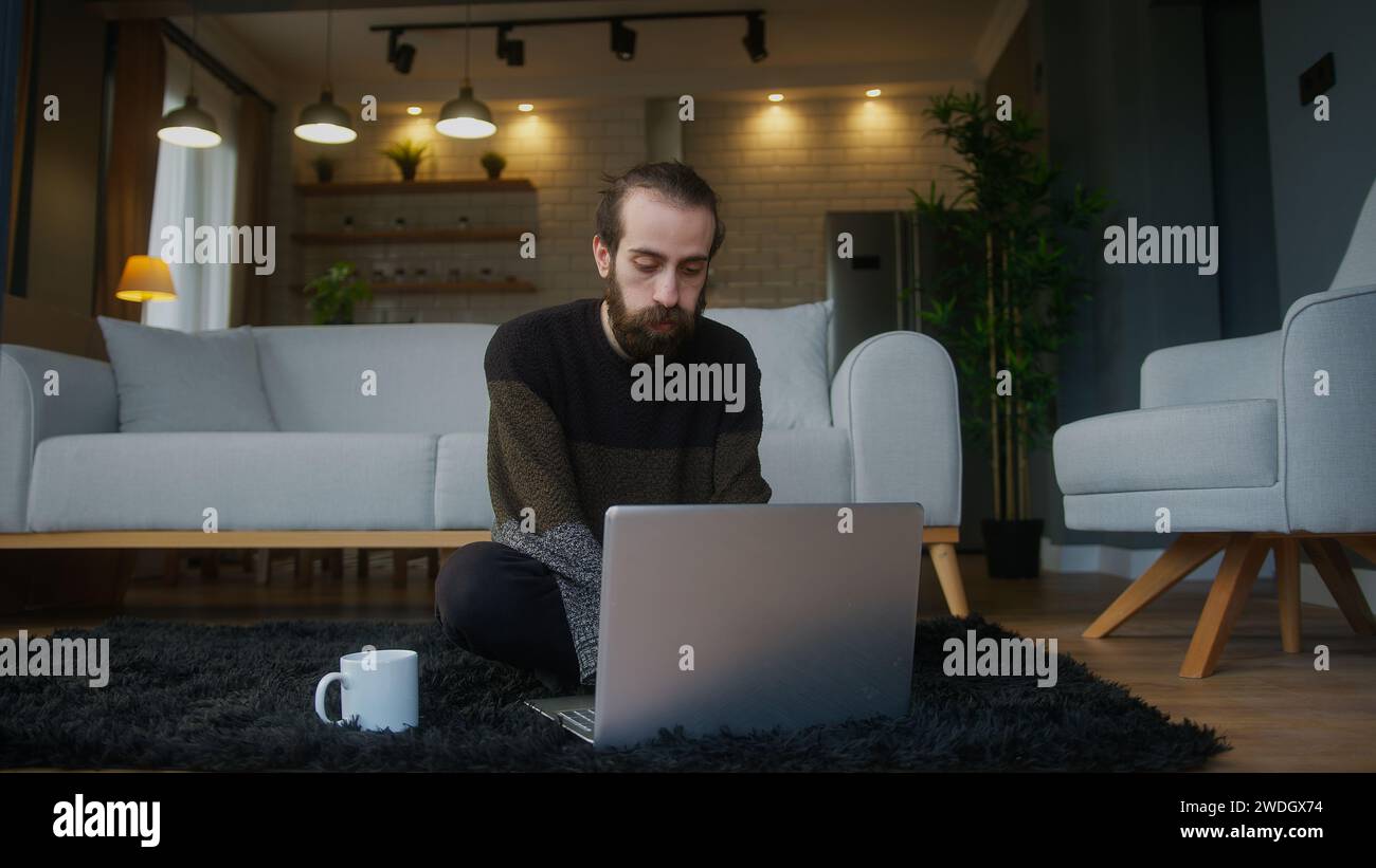 Gelangweilter junger bärtiger Mann, der auf dem Boden sitzt und einen Laptop benutzt, macht Fernarbeit Stockfoto