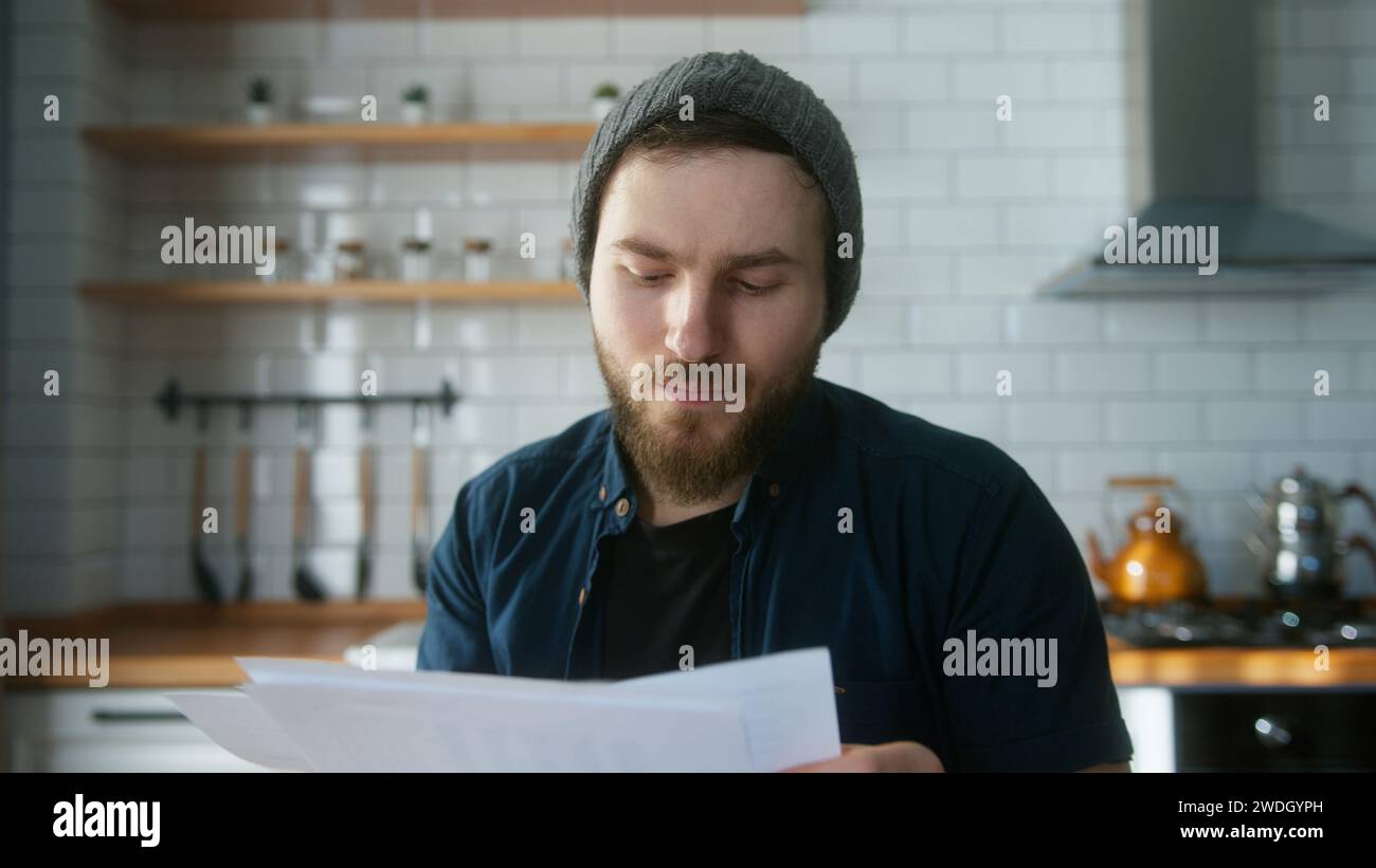 Porträt eines jungen erwachsenen Mannes mit Beanie, der in der modernen Küche zu Hause sitzt, spricht, die Berichte in Videokonferenzgesprächen ansieht. Selbstbewusste männliche haben Stockfoto