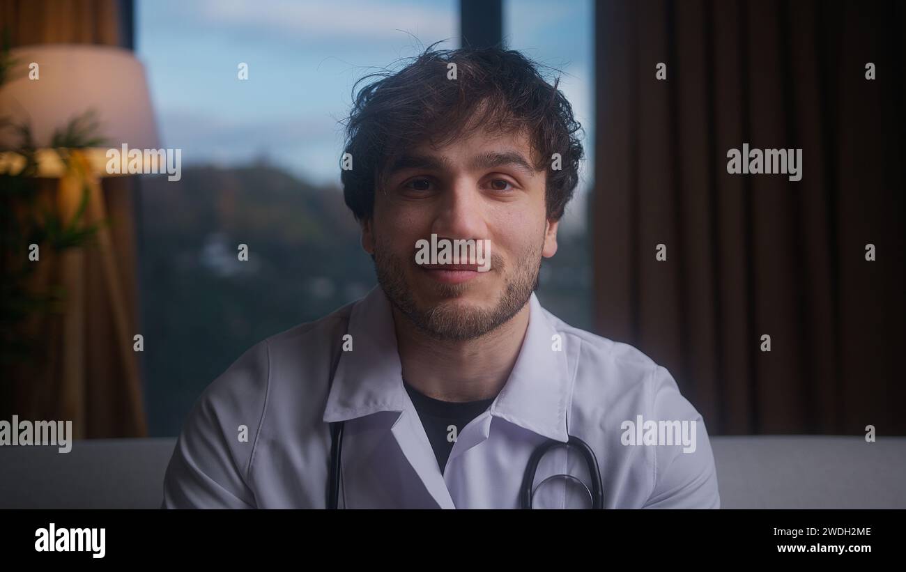 Lächelnder junger professioneller männlicher Doktor tragen weiße Uniform mit Stethoskop, der Videokonferenzen führt, online mit dem Patienten chatten und die Kamera hineinblickt Stockfoto