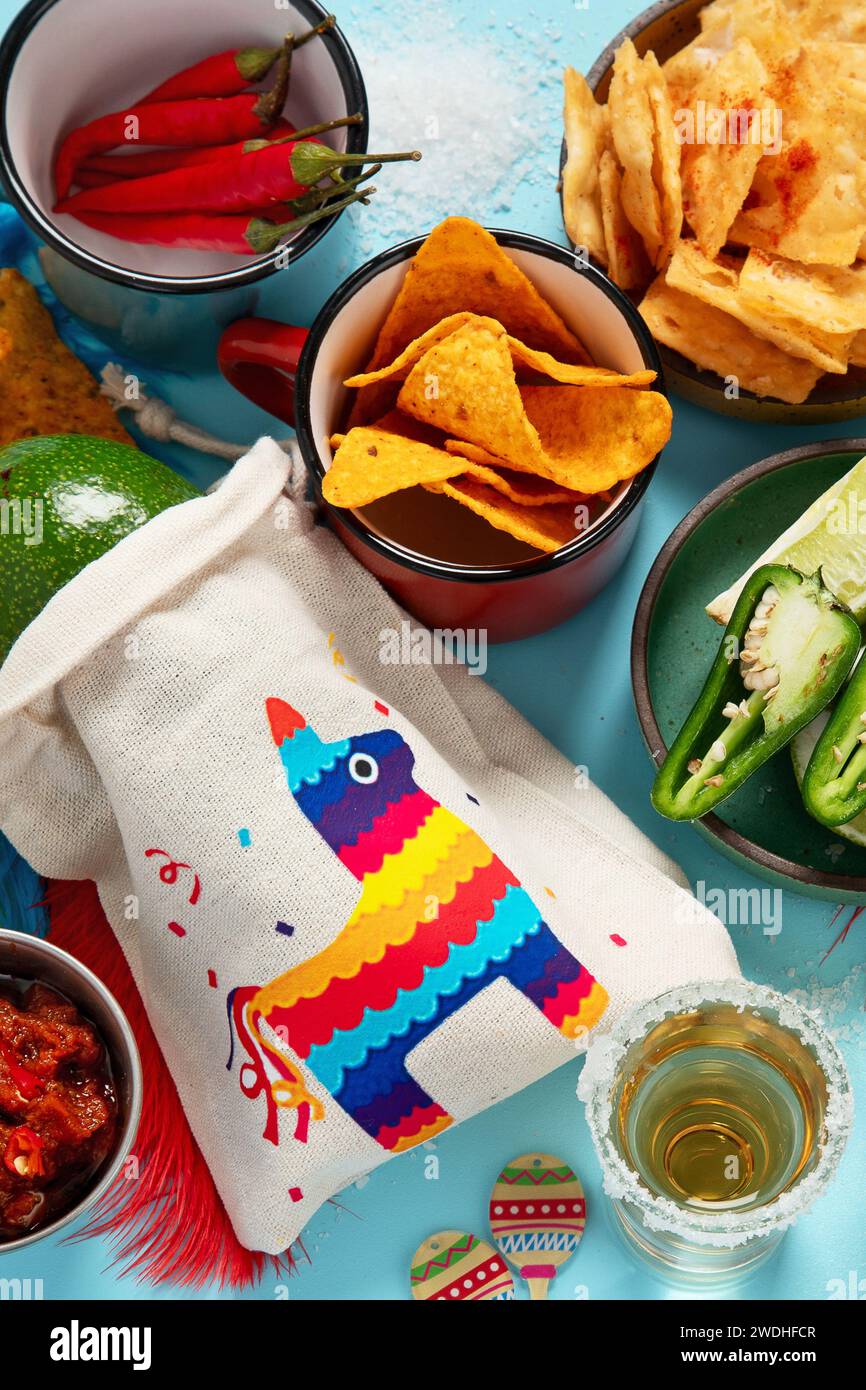 Cinco-de-Mayo-Konzept. Traditionelles Essen Nacho Chips Salsa Sauce Chili Tequila mit Salz Limette Sombrero Serape Kakteen und Maracas auf blauem Hintergrund. Stockfoto