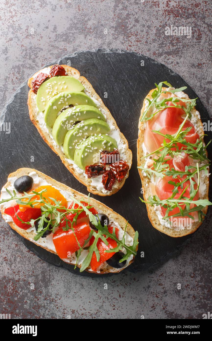 Gemischte Bruschetta-Sandwiches mit Rucola, Schinken, sonnengetrockneten Tomaten, Avocado, Frischkäse, Oliven und gebratene Paprika Nahaufnahme auf einem Schieferbrett auf t Stockfoto