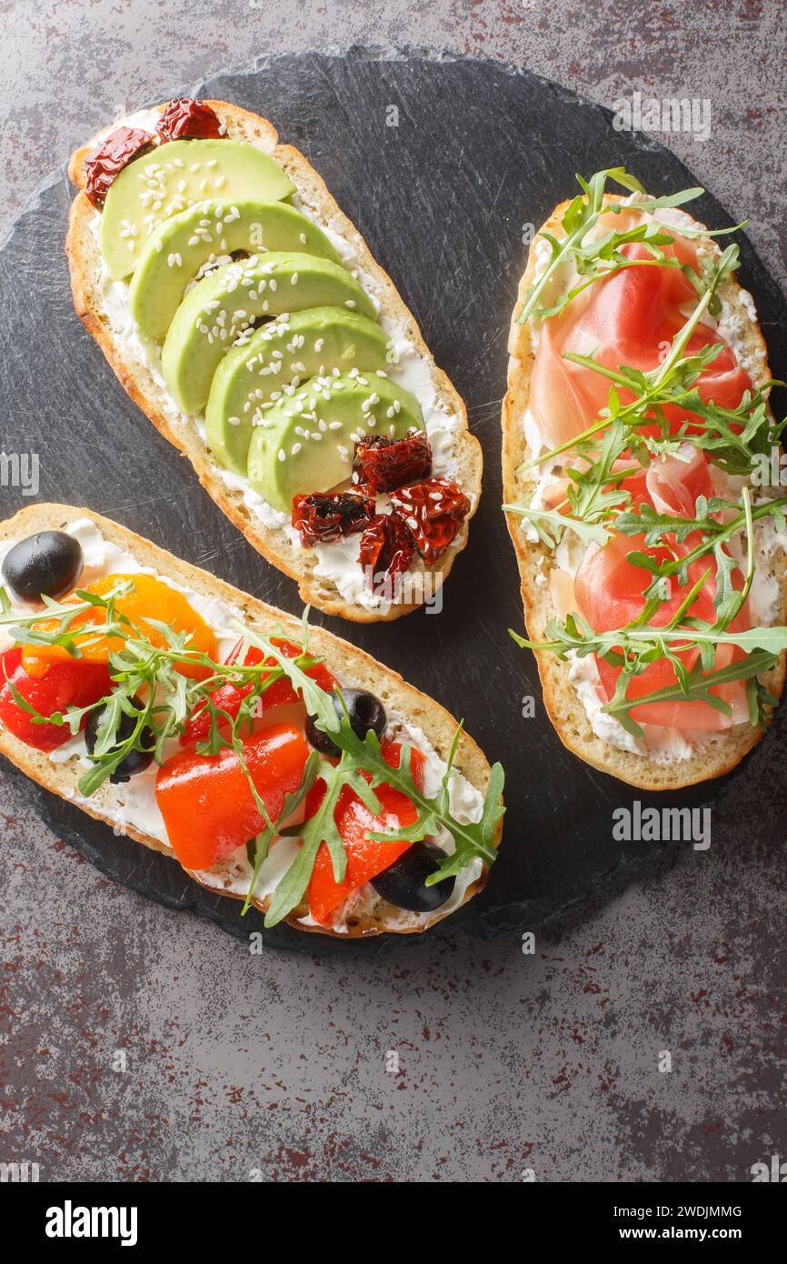Bruschetta-Sandwiches mit Rucola, Schinken, sonnengetrockneten Tomaten, Avocado, Frischkäse und gebratenen Paprika in Nahaufnahme auf einem Schieferbrett auf dem Tisch. Stockfoto