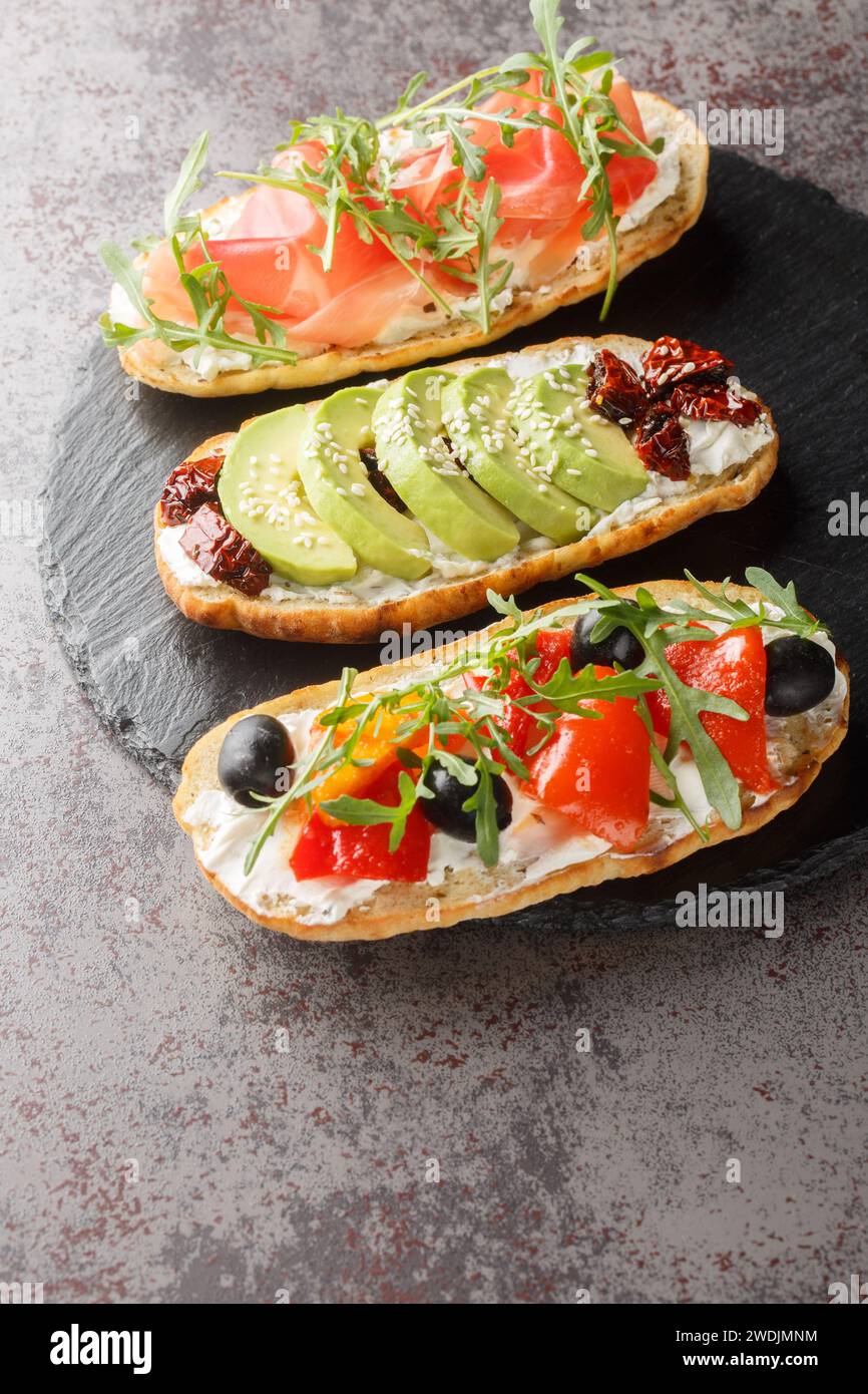 Bruschetta-Sandwiches mit Rucola, Schinken, sonnengetrockneten Tomaten, Avocado, Frischkäse und gebratenen Paprika in Nahaufnahme auf einem Schieferbrett auf dem Tisch. Stockfoto