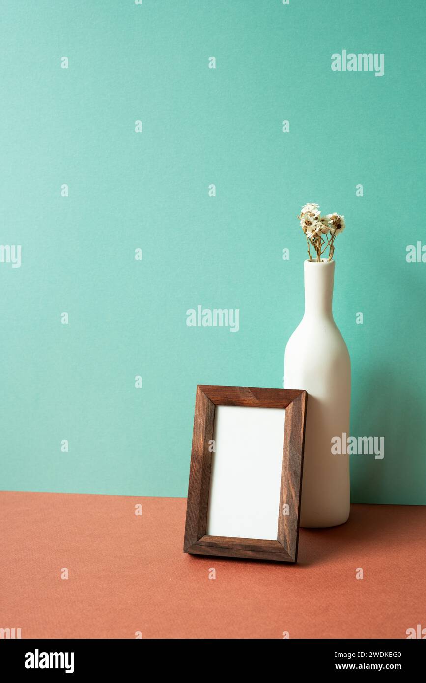Leerer hölzerner Bilderrahmen mit weißer Vase mit trockener Blume auf rotem Tisch. Mintgrüner Wandhintergrund Stockfoto