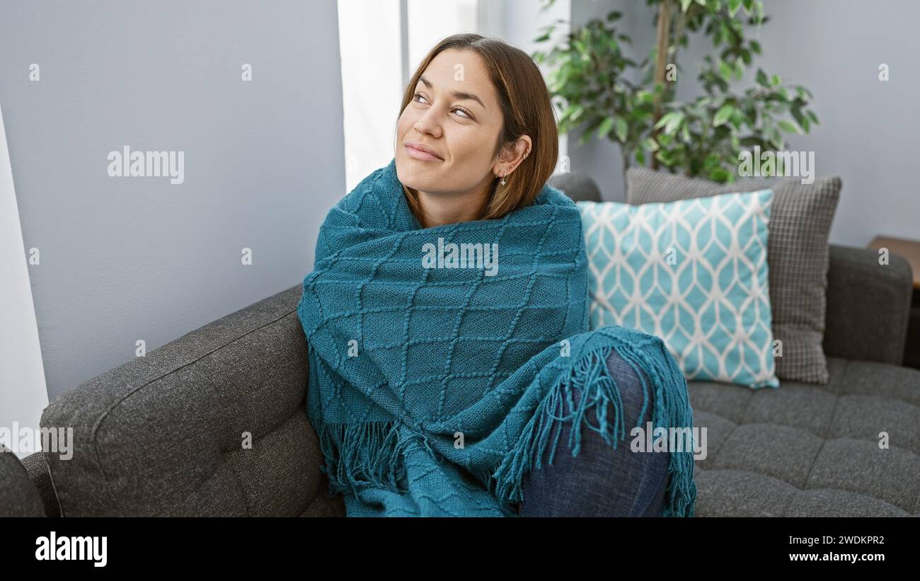Eine besinnliche junge Frau, in eine blaue Decke gewickelt, sitzt auf einem grauen Sofa in einem gemütlichen Wohnzimmer. Stockfoto Eine besinnliche junge Frau, in eine blaue Decke gewickelt, sitzt auf einem grauen Sofa in einem gemütlichen Wohnzimmer. Stockfoto
