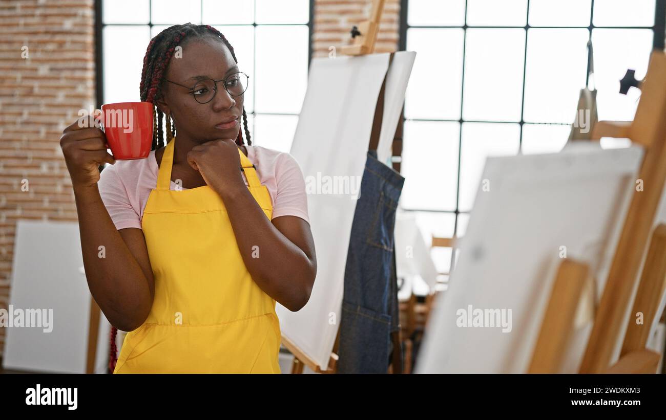 Ernsthafte aftere afroamerikanische Künstlerin, mit wunderschönen Zöpfen und Gläsern, mit einer Kaffeetasse und einem tiefen Gedanken über ihre nächste Zeichnung auf Leinwand im Stockfoto