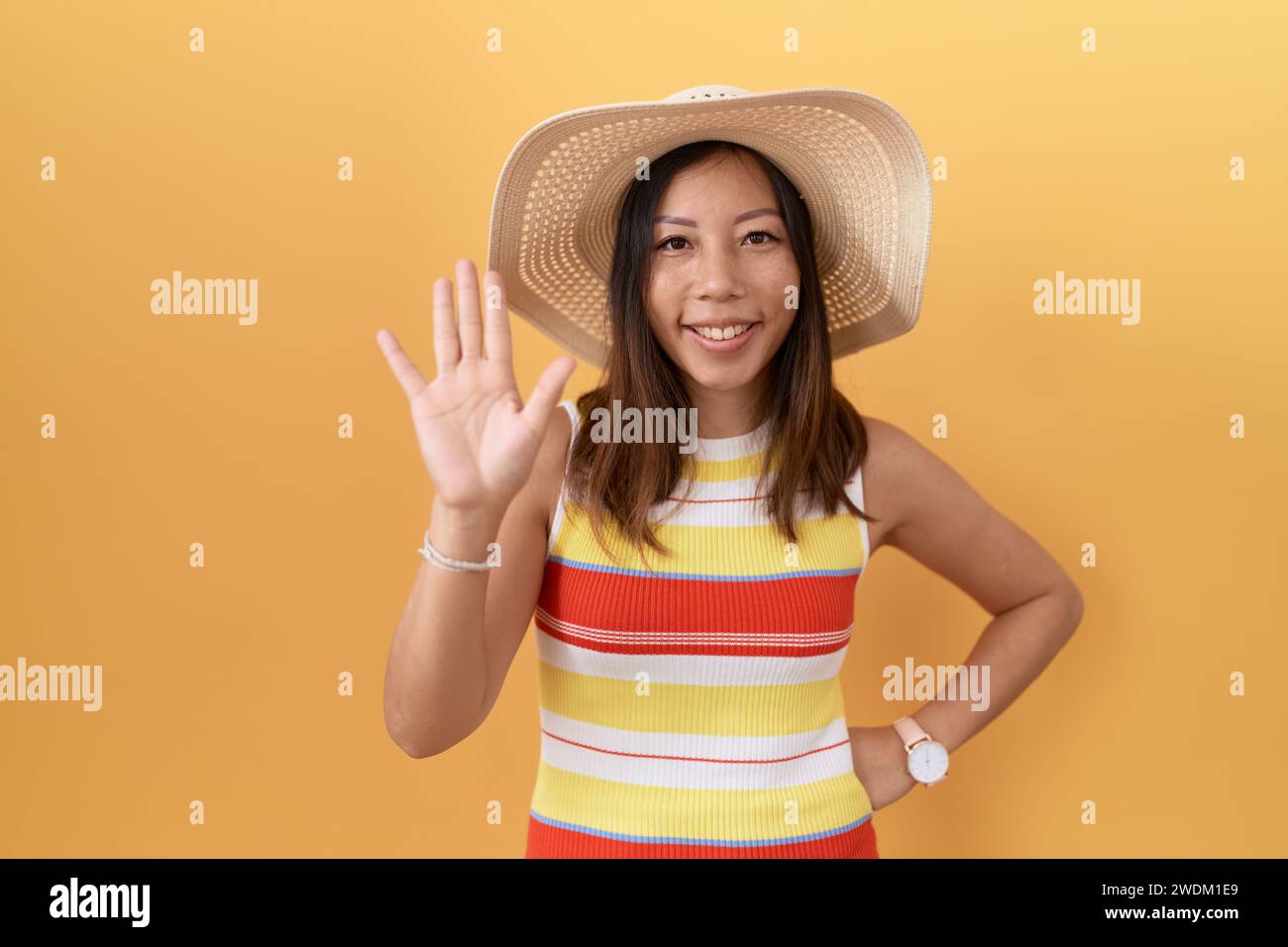 chinesin mittleren Alters, die einen Sommerhut über gelbem Hintergrund trägt, verzichtet darauf, Hallo zu sagen, glücklich und lächelnd, freundliche Willkommensgeste Stockfoto