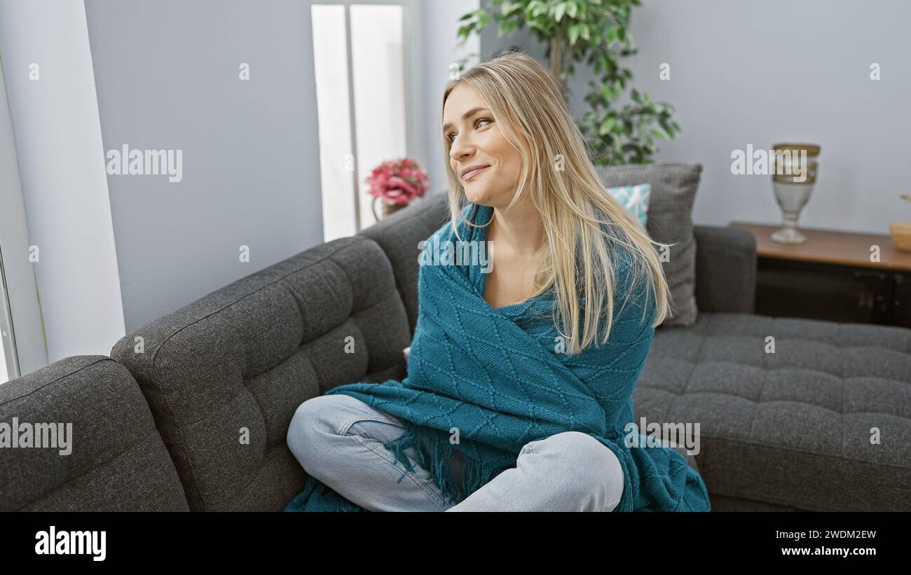 Eine chillige, junge blonde Frau, die sich in eine Decke kuschelt, sehnt sich nach Wärme und schaut durch ihr Fenster Stockfoto Eine chillige, junge blonde Frau, die sich in eine Decke kuschelt, sehnt sich nach Wärme und schaut durch ihr Fenster Stockfoto