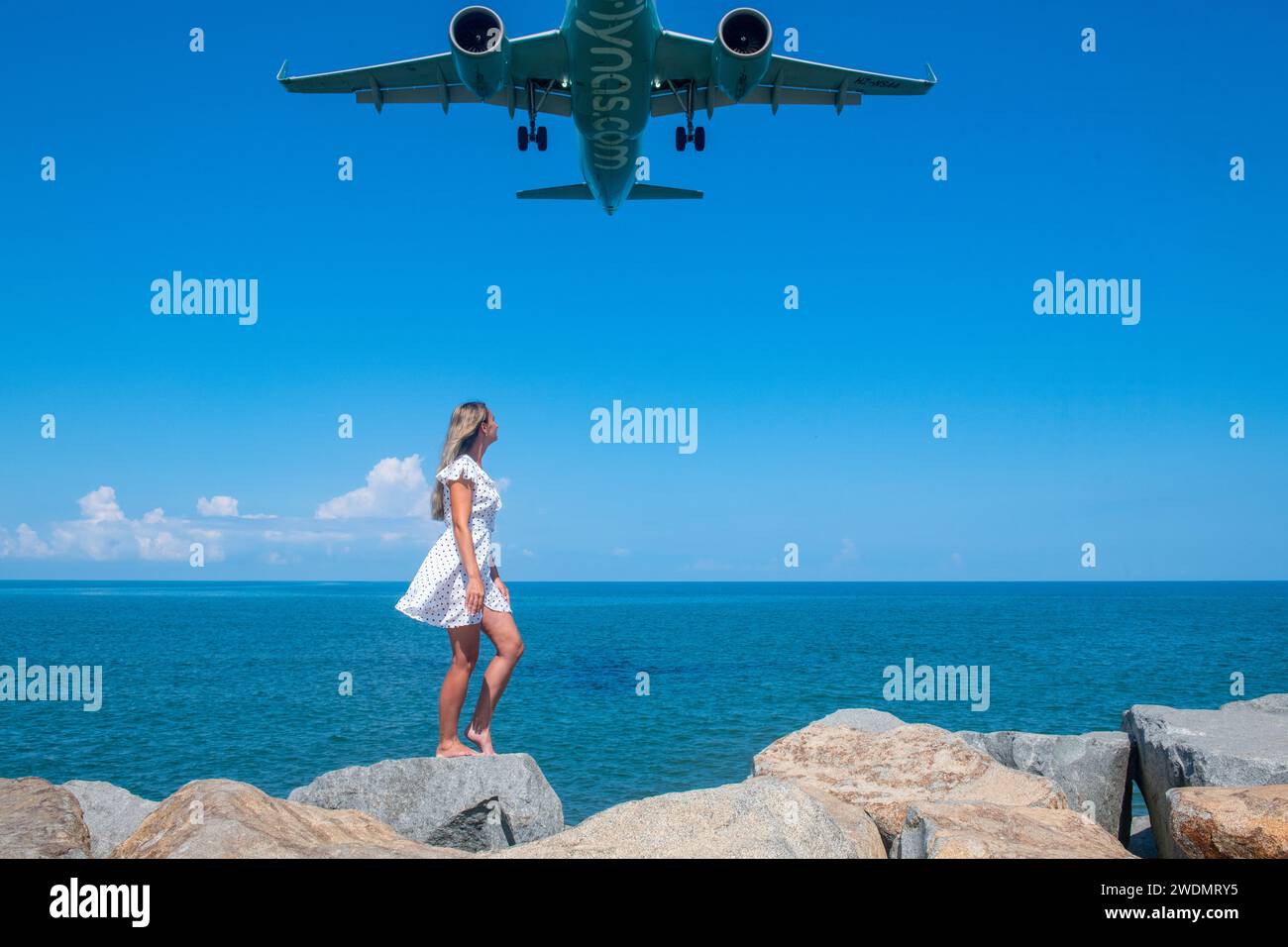 Coastal Encounter: Mädchen in weißem Kleid auf Steinen, ein Flugzeug, das vom Blauen Meer aus lockt Stockfoto