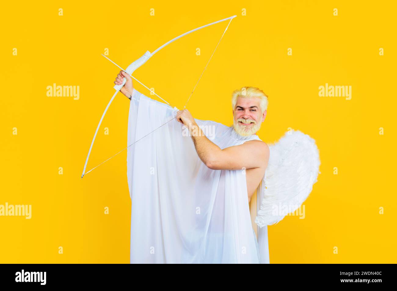 Valentinstag. Lächelnder bärtiger Mann im Engelskostüm, der mit Bogen zielt. Gott der Liebe. Valentinstag-Amulett in Engelsflügeln mit Bogen. Männlich Stockfoto