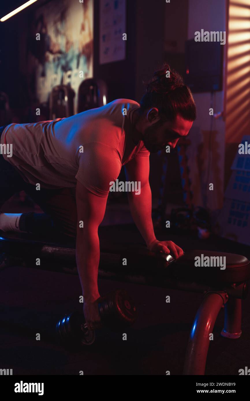 Konzentrierter männlicher Athlet, der sich in einem dynamischen Spiel aus blauen und roten Lichtern mit einem subtilen Nebel in der Luft an einer Kurzhantelreihenübung beteiligt. Stockfoto