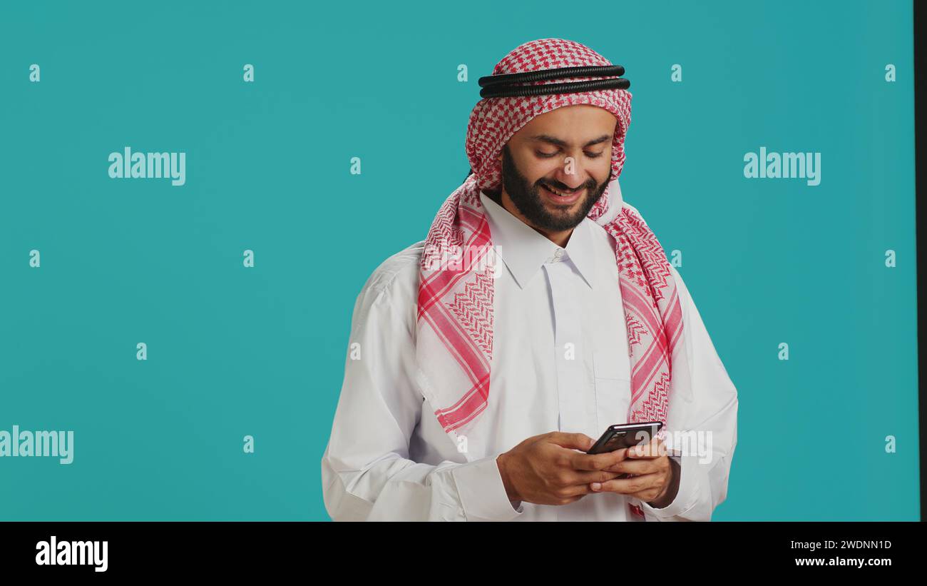 Lächelnder Erwachsener, der Nachrichten am Telefon schreibt und traditionelle muslimische Kleidung trägt. Islamisch-arabische Person in weiß gekleidet und karierte Gghutra, die auf Social-Media-Webseiten plaudert, SMS schreibt. Stockfoto