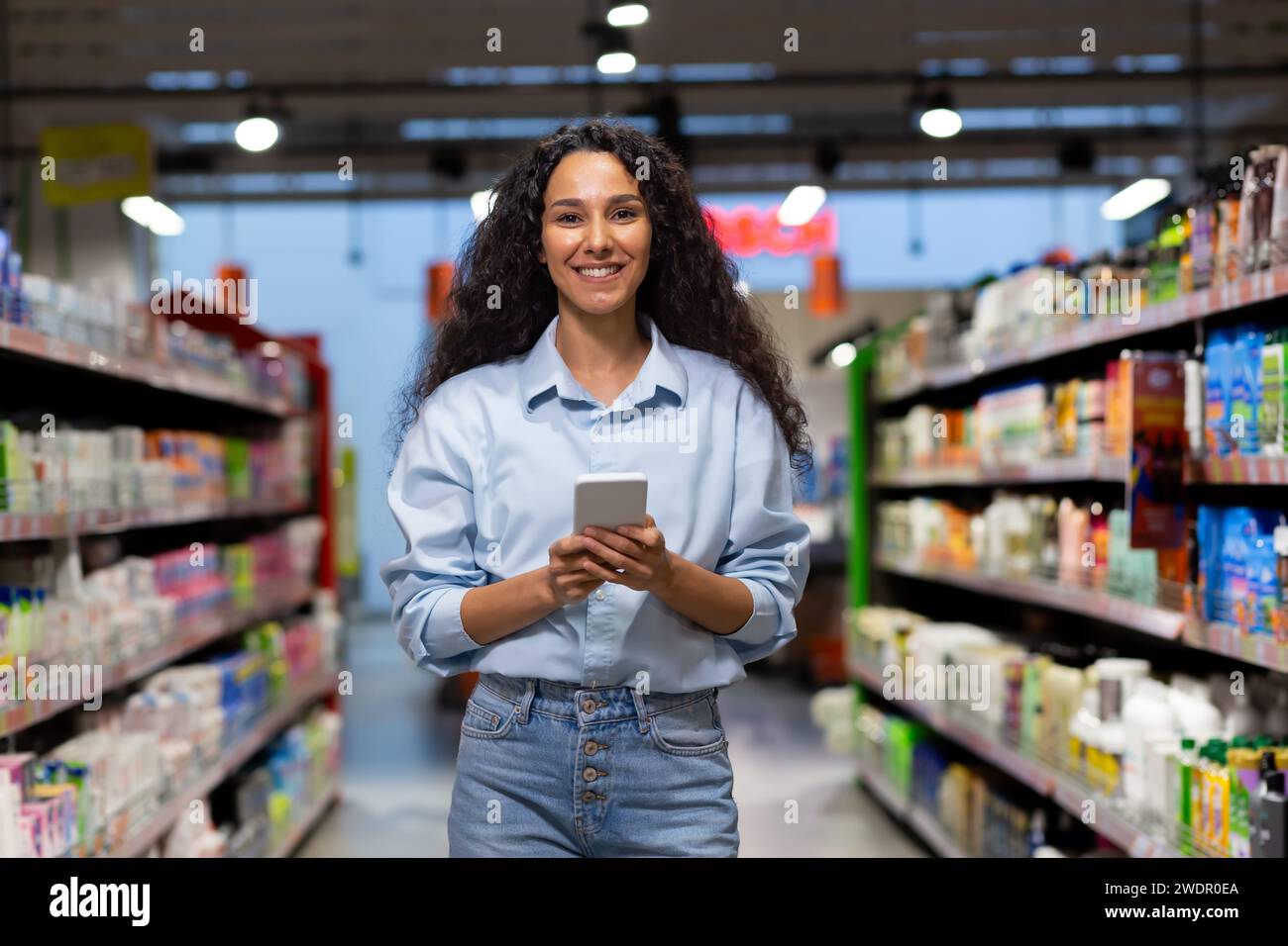 Lächelnde junge Käuferin, die ein Handy im Lebensmittelgeschäft benutzt, inmitten der farbenfrohen Regale voller Produkte. Stockfoto