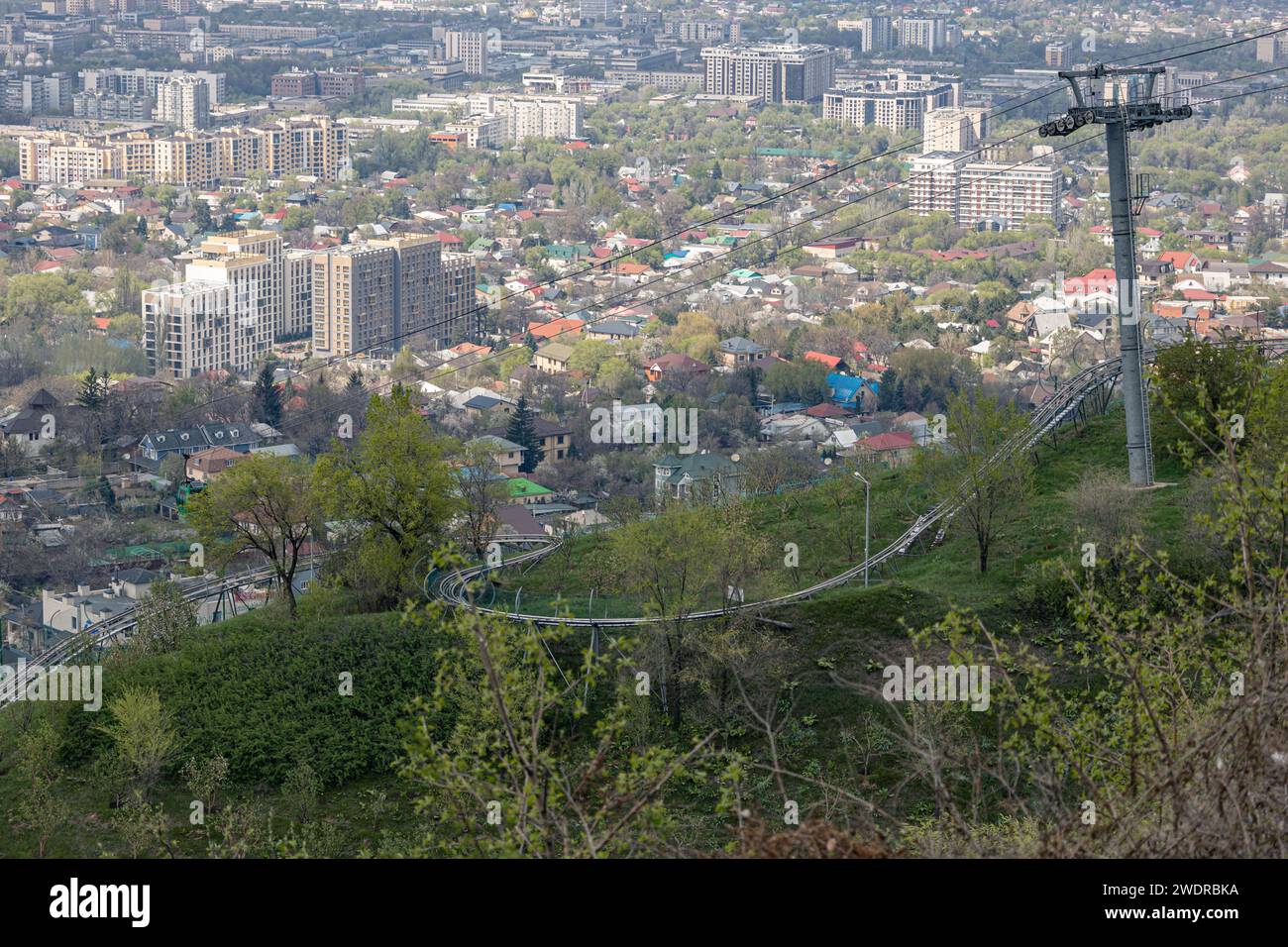 Leerer Sessellift führt zum Vergnügungspark Kok Tobe und der Rodelbahn am Berg. Grünes Gras und Büsche, sonniger Frühlingstag. Sehen Sie die Gebäude Almaty City Stockfoto