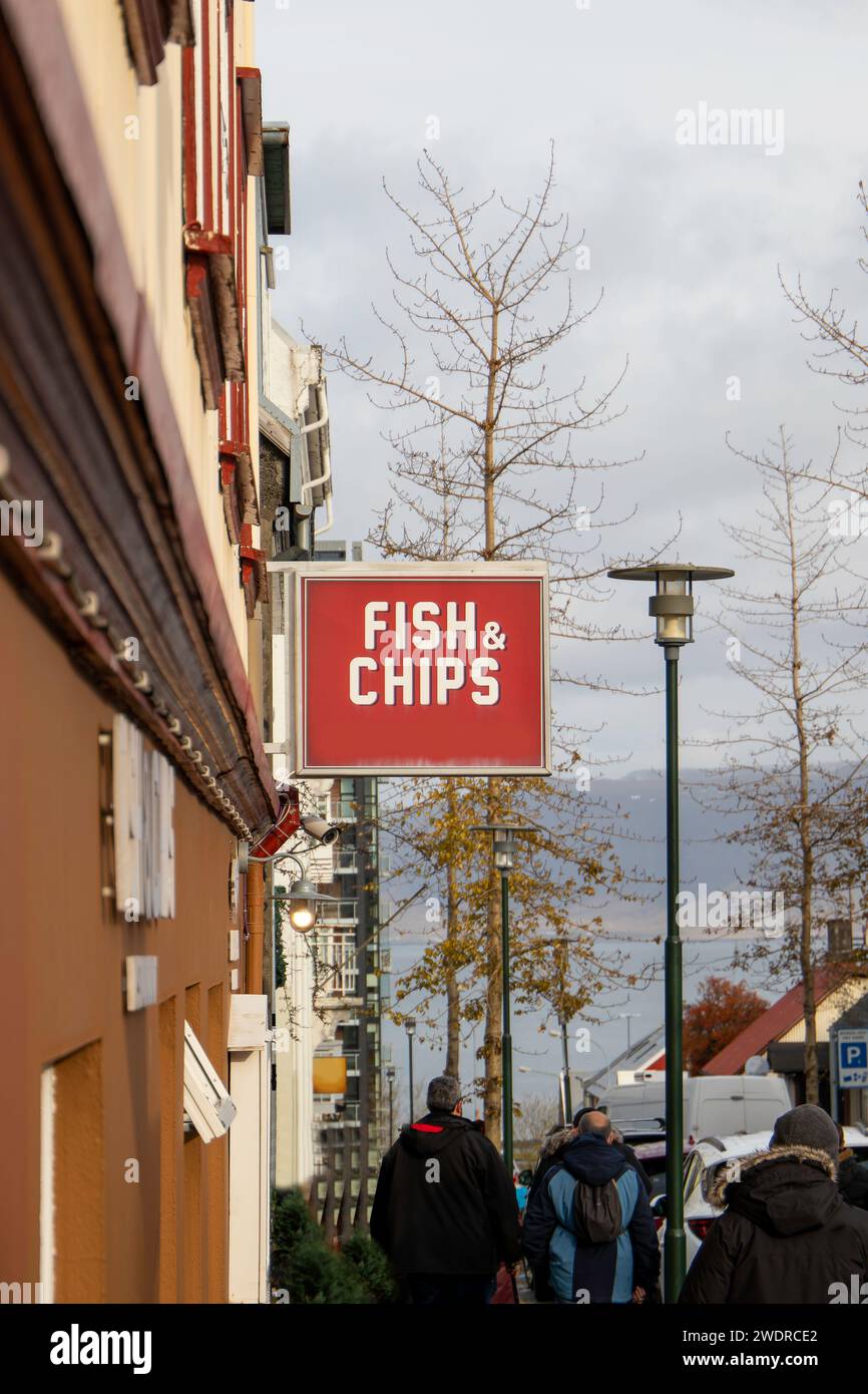 Rückansicht der anonymen Fußgänger in warmer Kleidung, während sie auf dem Bürgersteig in der Nähe des Gebäudes mit Restaurant, das Fish and Chips vor bedecktem Himmel serviert, spazieren gehen Stockfoto