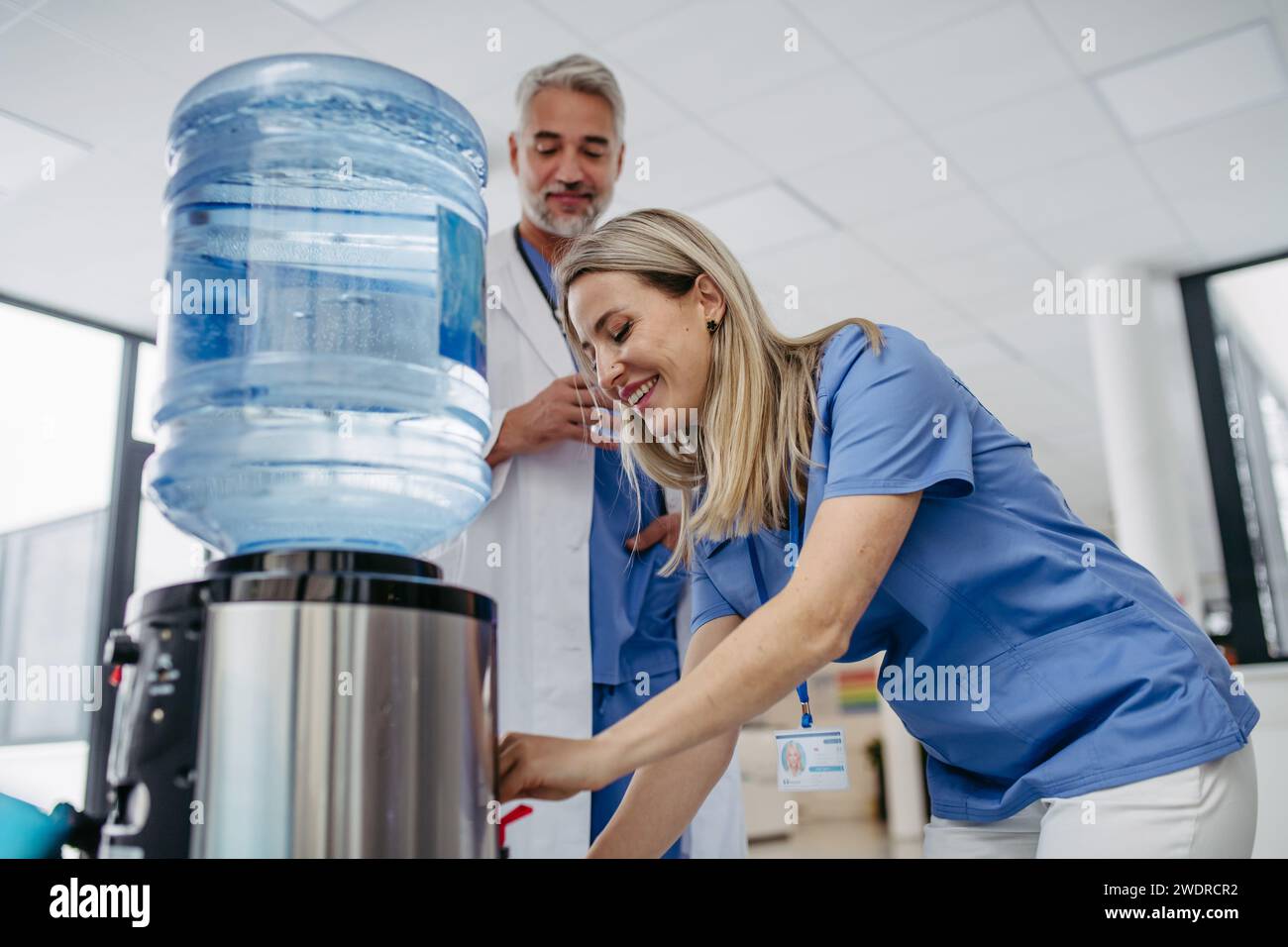 Arzt und Krankenschwester machen eine Pause während der Arbeitsschicht im Krankenhaus, trinken Wasser aus dem Wasserspender in der Krankenhauslobby, sprechen. Stockfoto