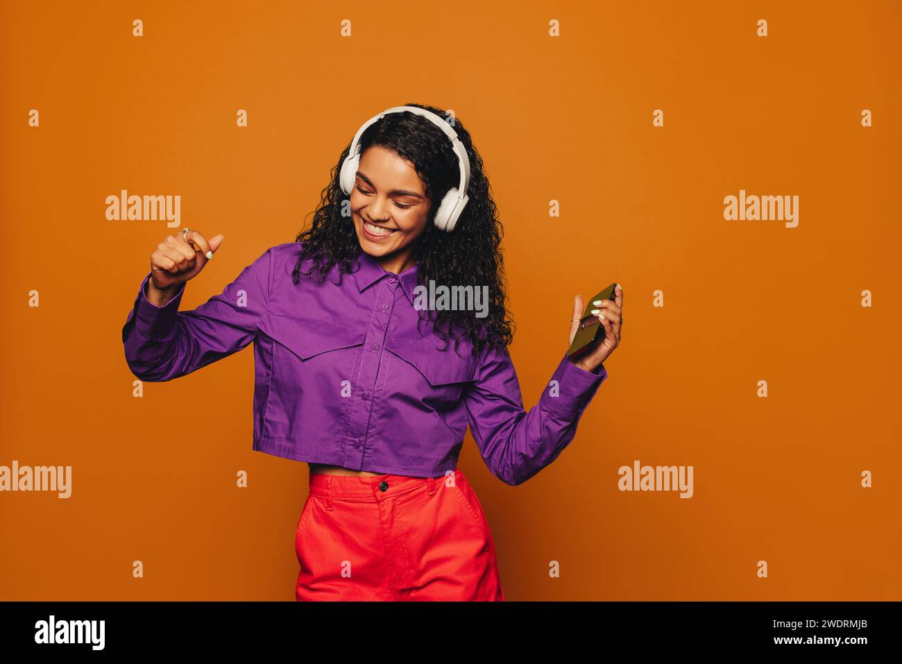 Auf orangefarbenem Hintergrund hält eine Frau in legerer Kleidung ein Telefon und hört Musik mit Kopfhörern. Ihr lockiges Haar hüpft, während sie zum bea tanzt Stockfoto