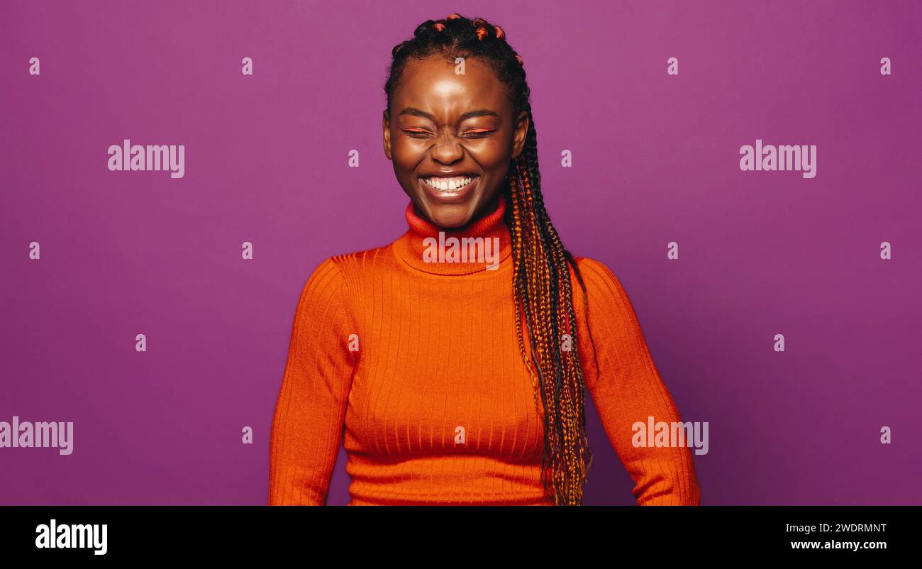 Selbstbewusste junge Frau mit stilvollen Zöpfen steht vor einem leuchtenden violetten Hintergrund, lächelt und strahlt Glück aus. Ihr modischer Gen Z-Stil und c Stockfoto