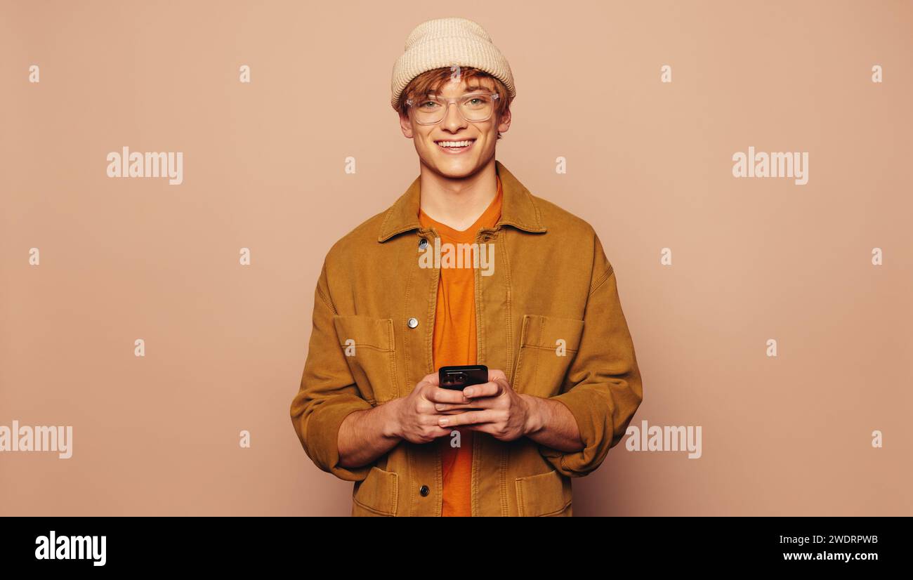 Happy man steht vor einem pfirsichfarbenen Hintergrund und hält ein Smartphone. Lässig gekleidet mit einer Jeansjacke und einer Brille, schaut er mit einer vibra in die Kamera Stockfoto
