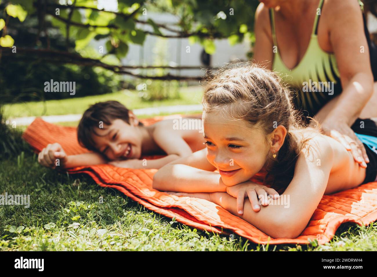 Oma beschmiert Kinder mit Sonnencreme, die auf dem Gras liegt Stockfoto