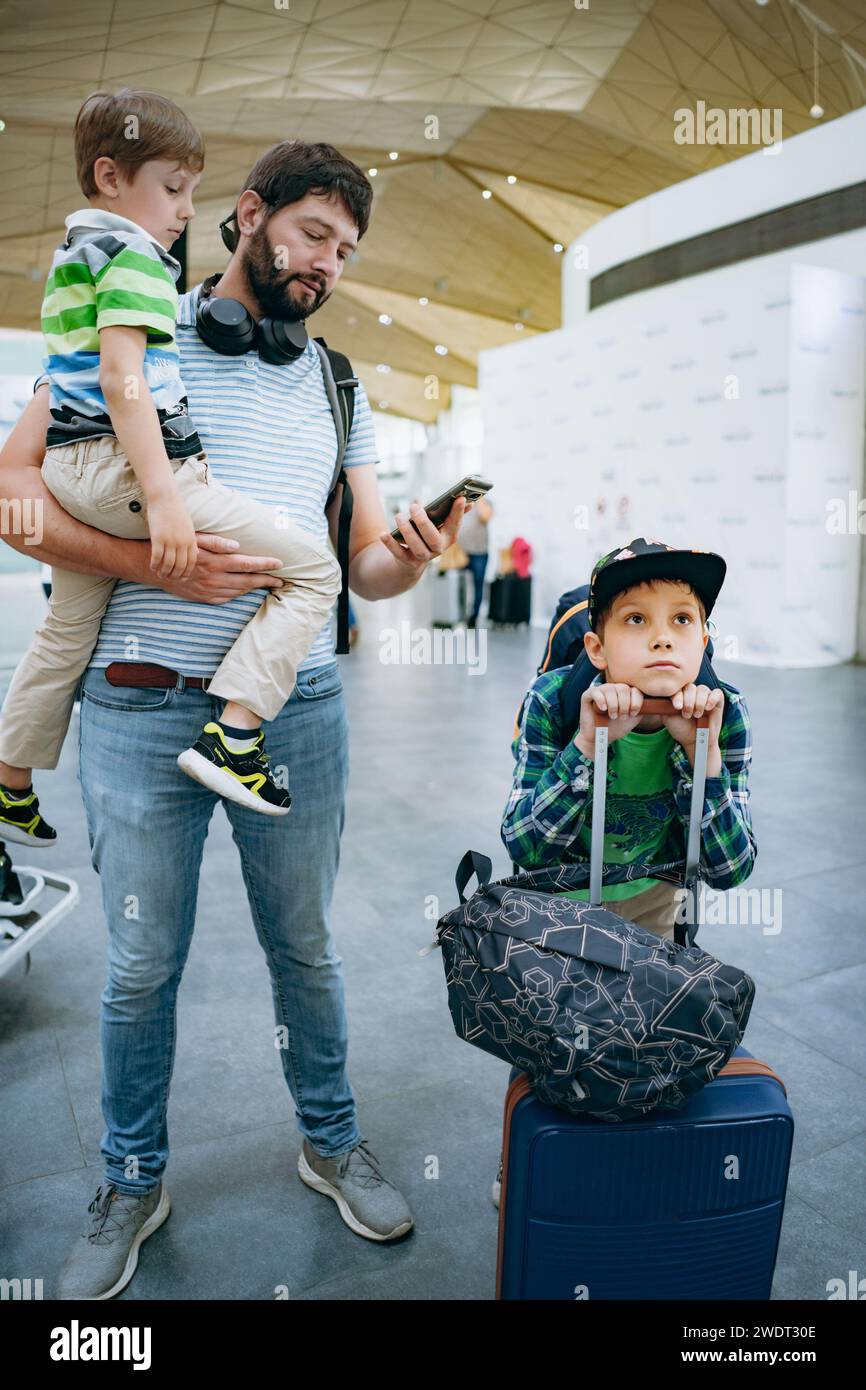 Vater hält süßen weißen Jungen am Flughafen. Kind mit Koffer und Rucksack wartet auf verspäteten Flug. Reisen mit Kindern Stockfoto