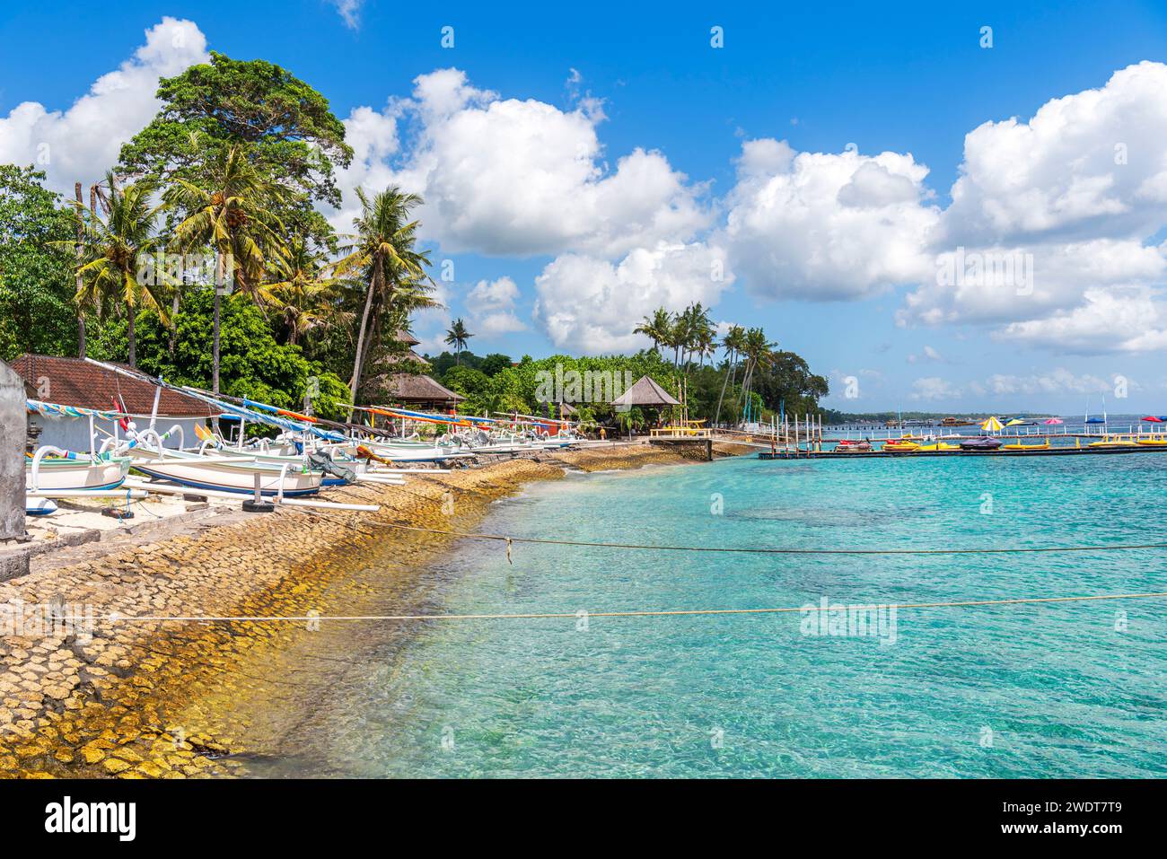 Blick auf einen tropischen Strand mit Palmen und Bäumen entlang der Küste der Insel Nusa Penida, mit traditionellen indonesischen Holzbooten Stockfoto