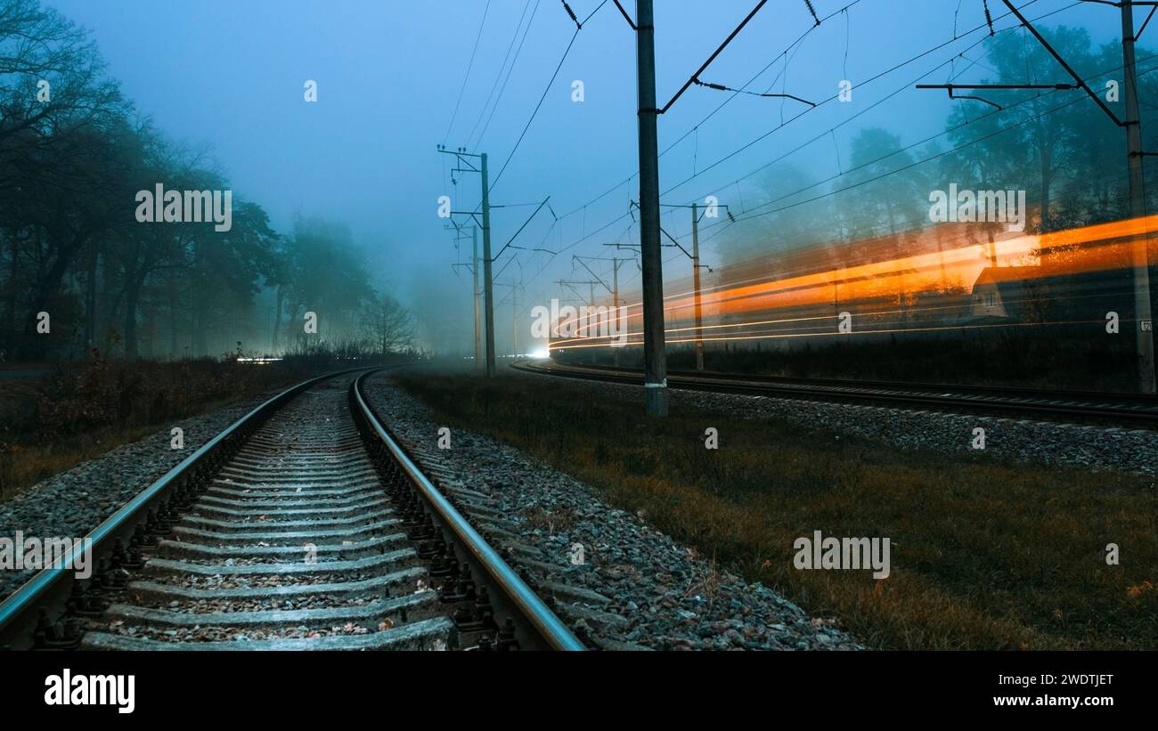 Die Eisenbahn an einem nebeligen Abend. Verschwommene Lichter eines vorbeifahrenden Zuges. Schnell vorbeifahrender Zug. Düster kalter Abend. Stockfoto