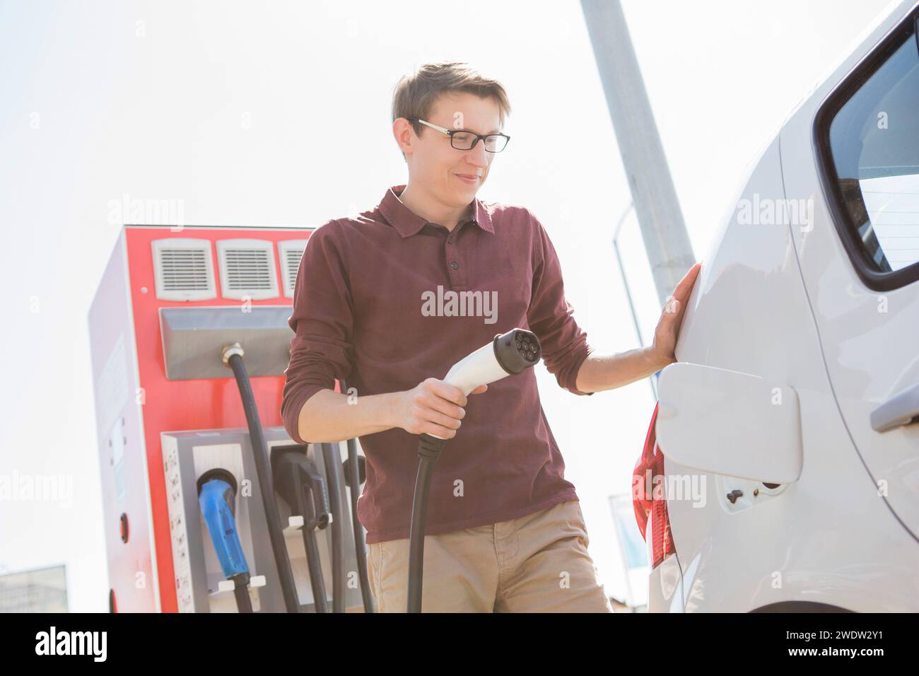 Ein Mann steht an der Ladestation und hält einen Stecker des Ladegeräts für ein Elektroauto. Stockfoto Ein Mann steht an der Ladestation und hält einen Stecker des Ladegeräts für ein Elektroauto. Stockfoto