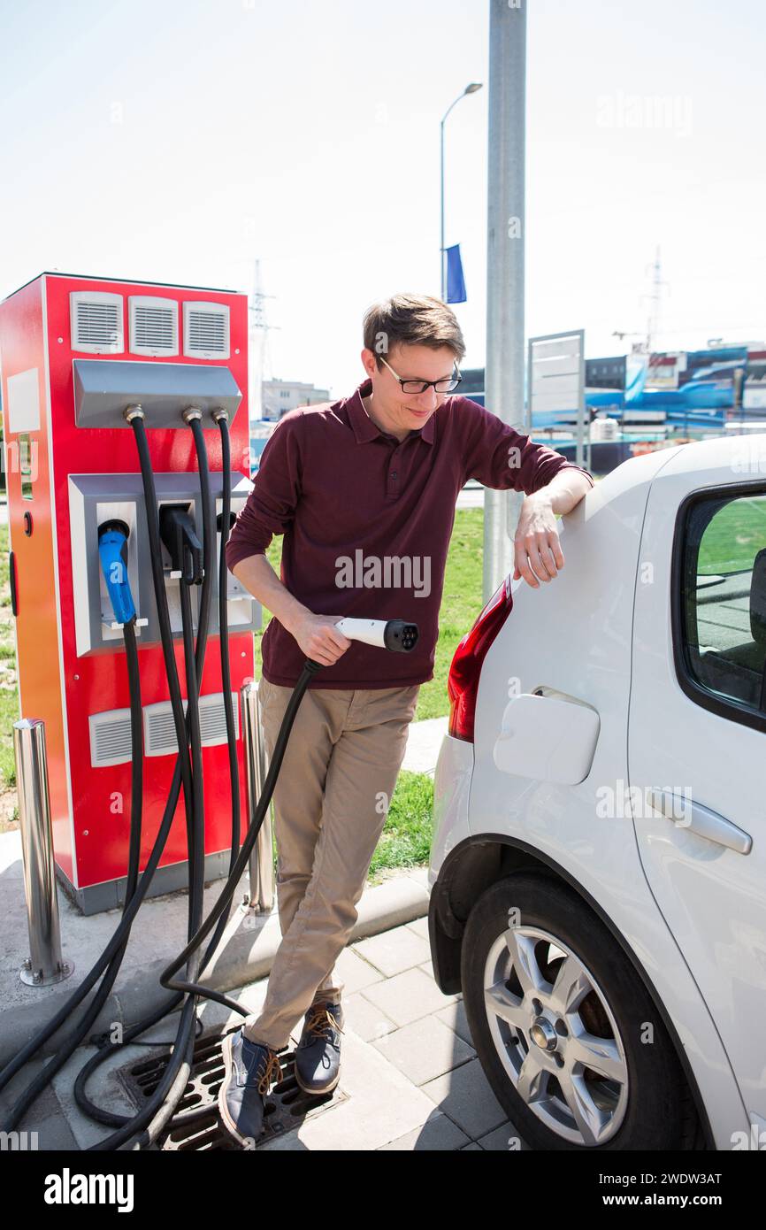 Ein Mann steht an der Ladestation und hält einen Stecker des Ladegeräts für ein Elektroauto. Stockfoto Ein Mann steht an der Ladestation und hält einen Stecker des Ladegeräts für ein Elektroauto. Stockfoto