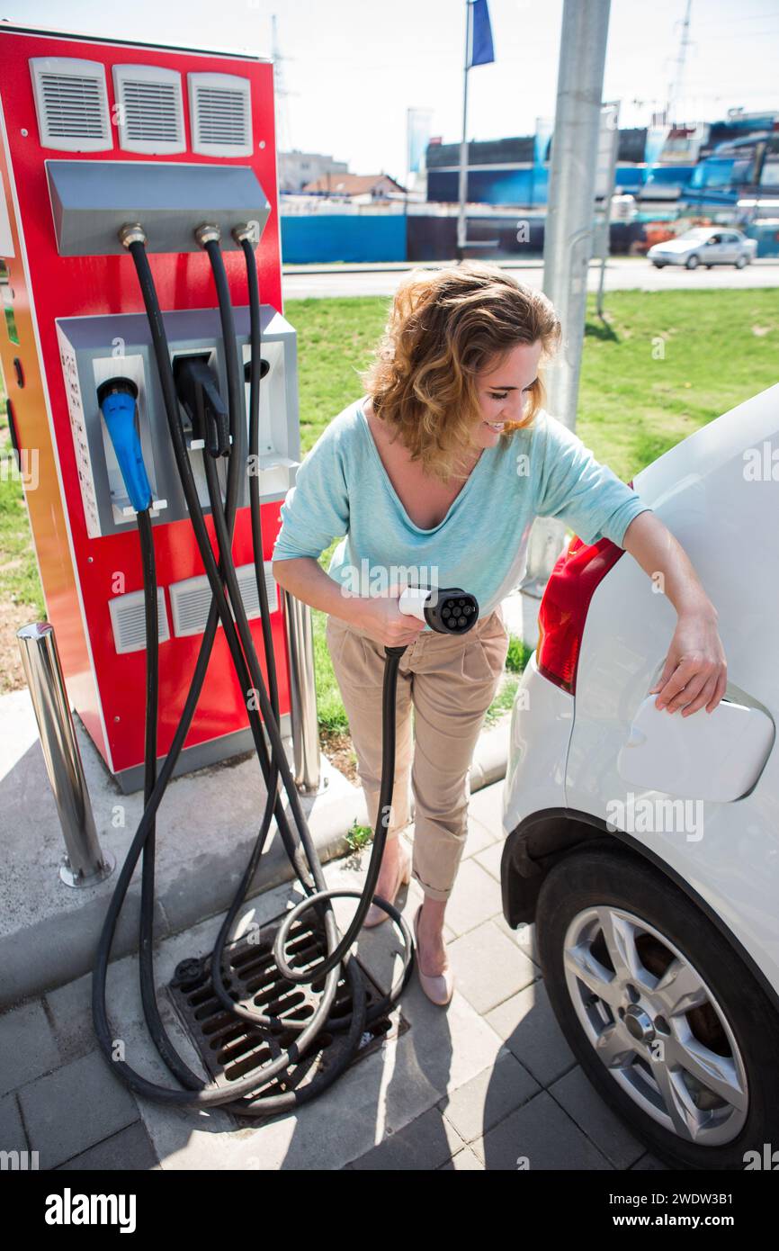 Eine Frau steht an der Ladestation und hält den Stecker des Ladegeräts für ein Elektroauto. Stockfoto Eine Frau steht an der Ladestation und hält den Stecker des Ladegeräts für ein Elektroauto. Stockfoto