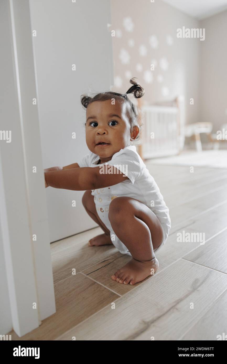Gemischtes Kleinkind spielt und versteckt sich hinter der geöffneten Tür im Zimmer. Afrokaukasisches kleines Mädchen. Das Konzept der interrassischen Familie. Stockfoto