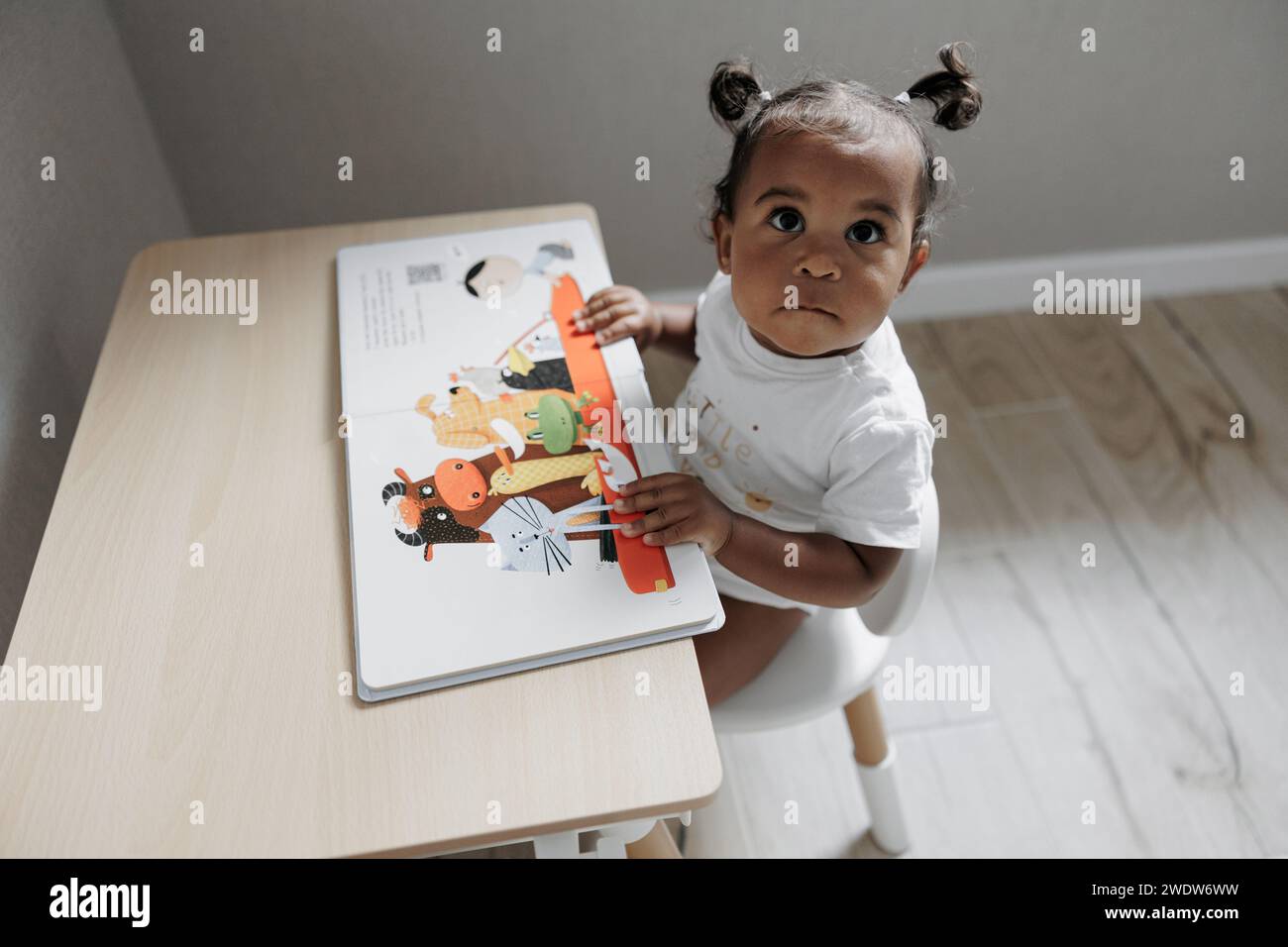 Gemischte Rasse Kleinkind Mädchen sitzt im Kinderzimmer neben dem Kindertisch und sieht Bilder im Kinderbuch. Afrokaukasisches kleines Mädchen. Das Konzept der Interracia Stockfoto