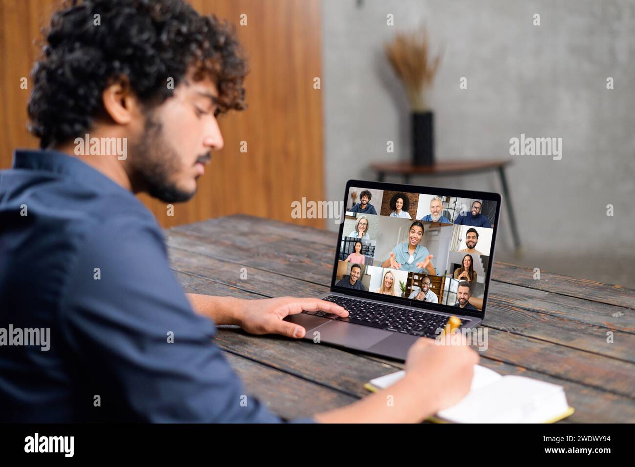 Ein konzentrierter indischer Mann sitzt an einem hölzernen Schreibtisch und nimmt an einer Videokonferenz auf seinem Laptop Teil. Er ist bereit, wichtige Punkte zu notieren und die interaktive Natur der Fernkommunikation zu demonstrieren Stockfoto