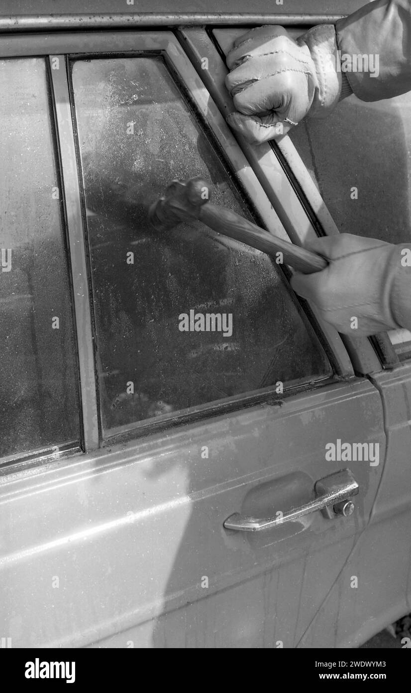 1970er Jahre, historisch, ein Handdieb mit Handschuhen, der versucht, ein Autofenster zu brechen, ein hinteres Lüftungsfenster, möglicherweise ein Ford Cortina MKII Estate Car, mit einem Hammer, England, Großbritannien. Stockfoto