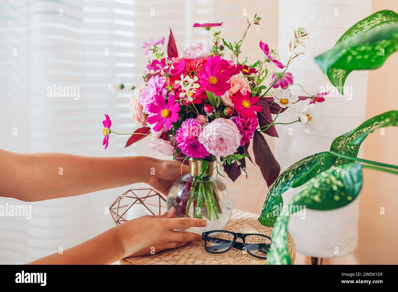 Die Frau legt einen Strauß rosa Blumen in eine transparente Vase auf den Rattan-Pad-Tisch. Die Behaglichkeit des Innenraums und die Sommerdekoration zu Hause werden gepflegt. Nahaufnahme Stockfoto