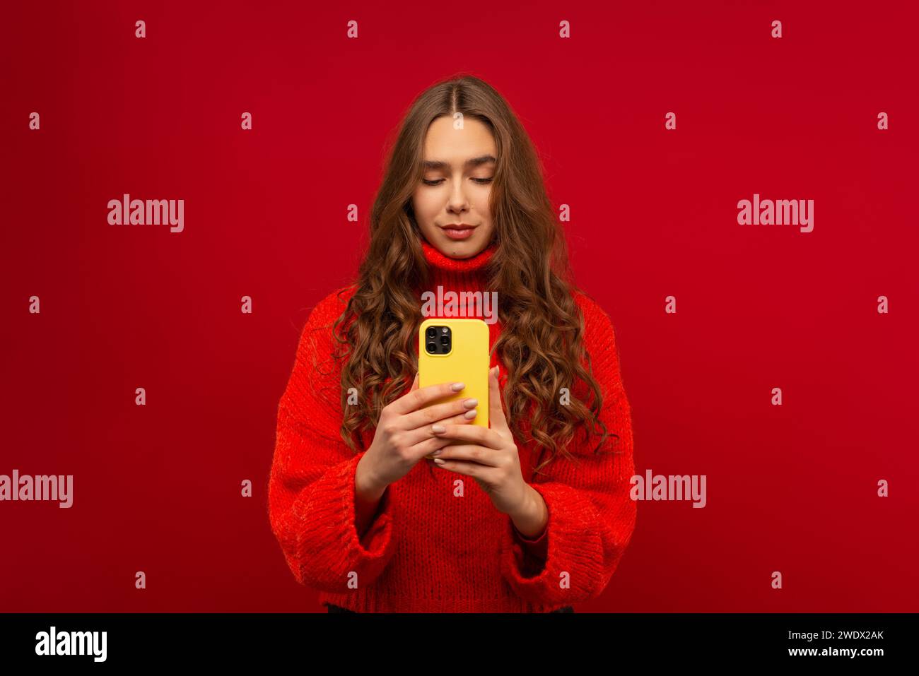 Porträt einer lächelnden emotionalen jungen Frau in lockigem Haar Generation Z in einem roten Pullover mit einem modernen Smartphone in einem Studio mit rotem Hintergrund Stockfoto