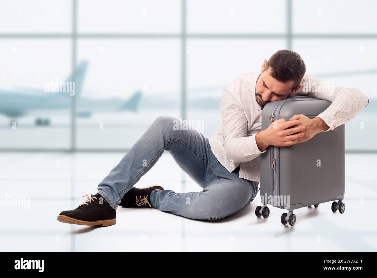 Bärtiger Mann, der auf dem Boden sitzt und seine Reisetasche umarmt, schläft und müde ist, auf einem Flughafen zu warten. Lange Warteschlangen. Flugverspätungen. Die Verzweiflung ist vorbei Stockfoto