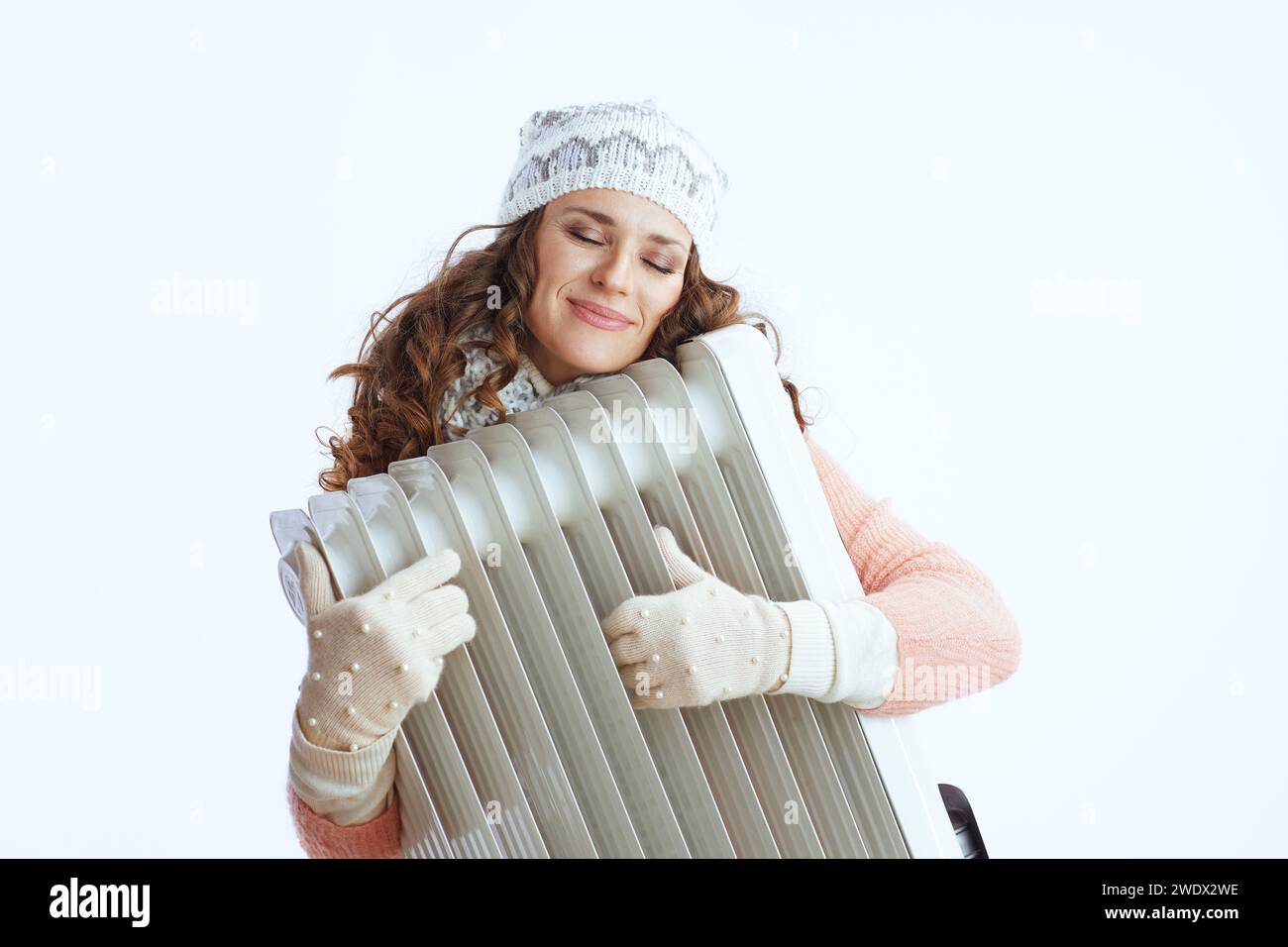 Hallo Winter. Entspannte stilvolle Frau im Pullover, Fäustlingen, Hut und Schal isoliert auf weißem Hintergrund mit Heizung. Stockfoto