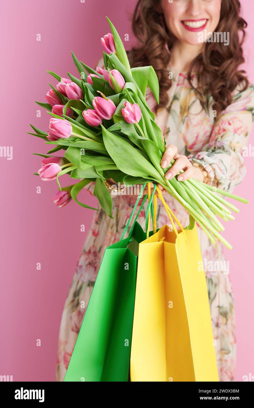 Nahaufnahme einer lächelnden Frau mittleren Alters im Blumenkleid mit Tulpenstrauß und Einkaufstaschen isoliert auf rosa Hintergrund. Stockfoto