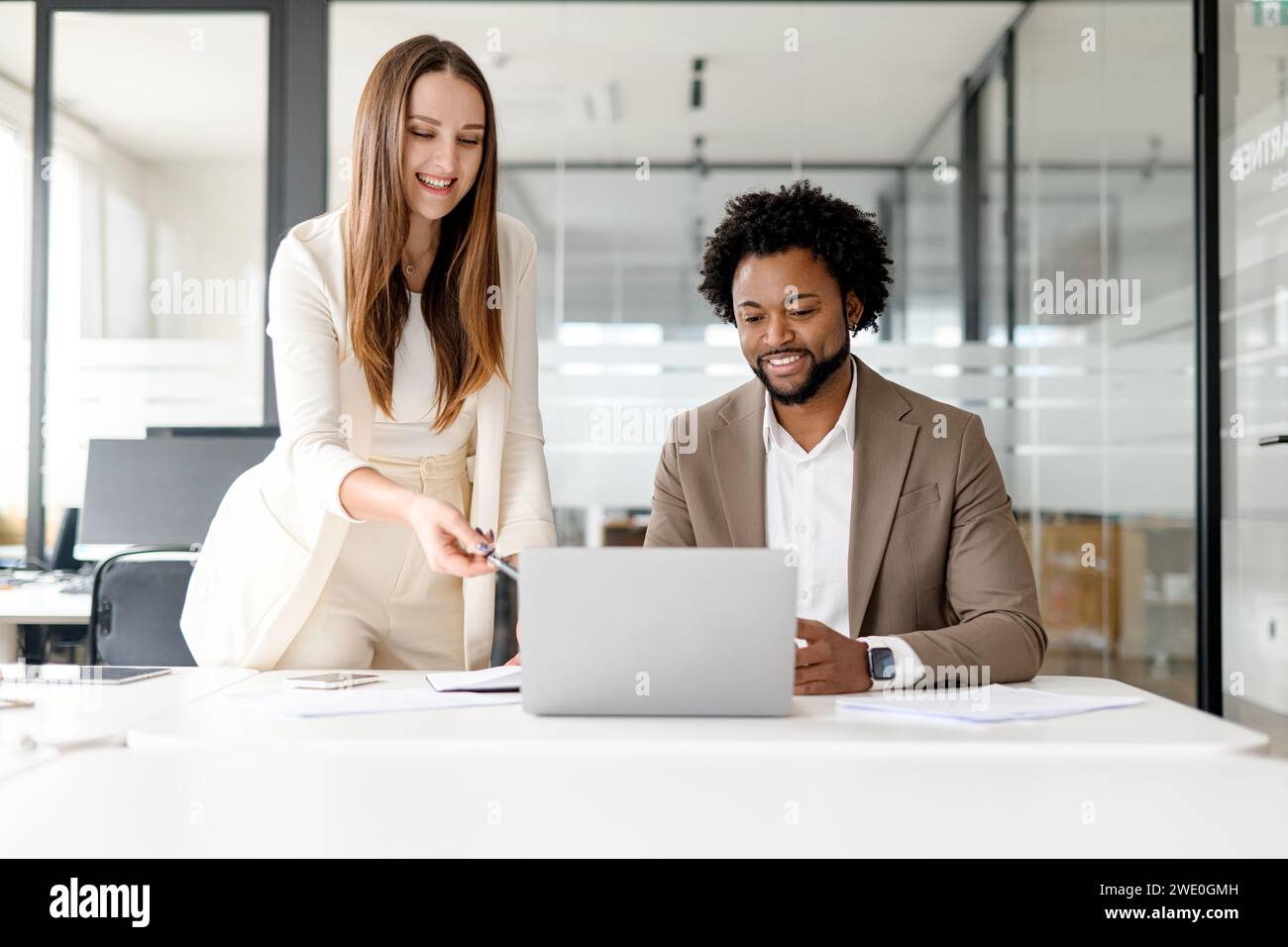Eine Frau zeigt auf einen Laptop-Bildschirm, während sie mit einem männlichen Kollegen über Inhalte spricht und interaktive Kommunikation und Teamarbeit in einem professionellen Kontext veranschaulicht. Stockfoto