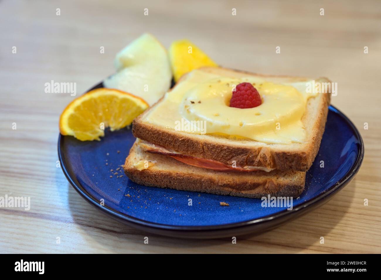 Toast Hawaii, gegrilltes offenes Sandwich mit Schinken, Ananas und Käse, ein paar Obststückchen und Himbeergarnitur auf einem blauen Teller auf einem Holztisch, Sel Stockfoto
