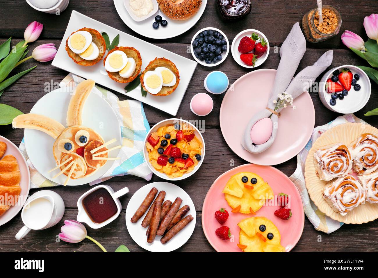 Lustiges Osterfrühstück oder Brunch-Tisch. Ansicht von oben nach unten auf dunklem Holzhintergrund. Hasenpfannkuchen, Eiernester, Hühnerfrüchte und verschiedene Frühlingsspeisen Stockfoto