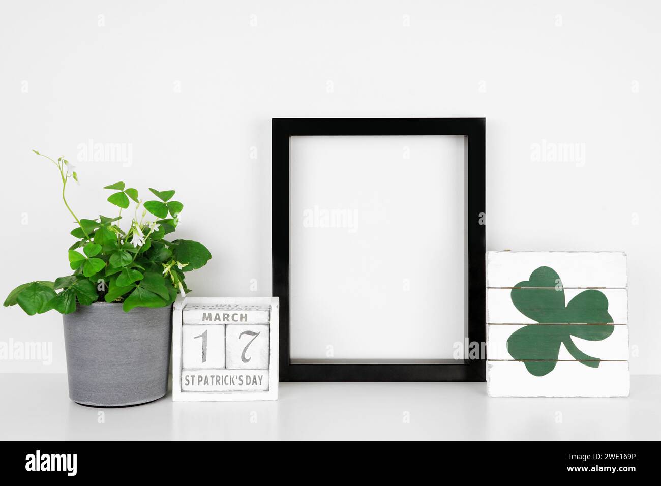 Schwarzer Rahmen mit St. Patricks Day-Dekor auf weißem Regal. Shamrock-Pflanze, schäbiger, schicker Holzkalender und Schild. Portraitrahmen gegen eine weiße Wa Stockfoto
