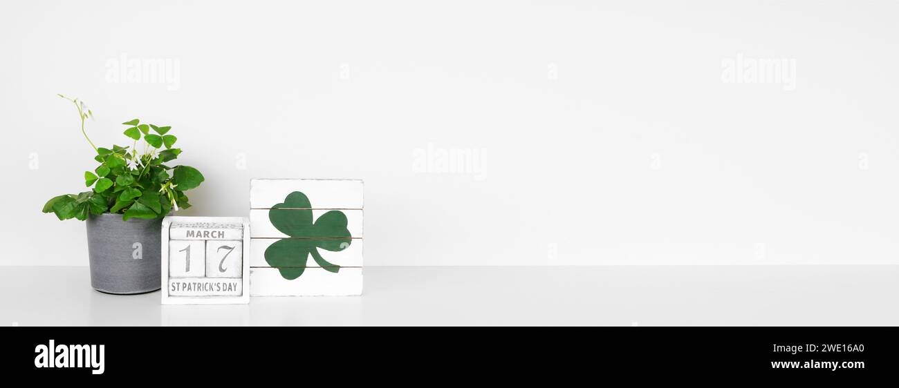 St. Patricks Day Dekor auf weißem Regal. Shamrock Pflanze, schäbiger schicker Holzkalender und Schild vor einem weißen Wandbanner. Kopierbereich. Stockfoto