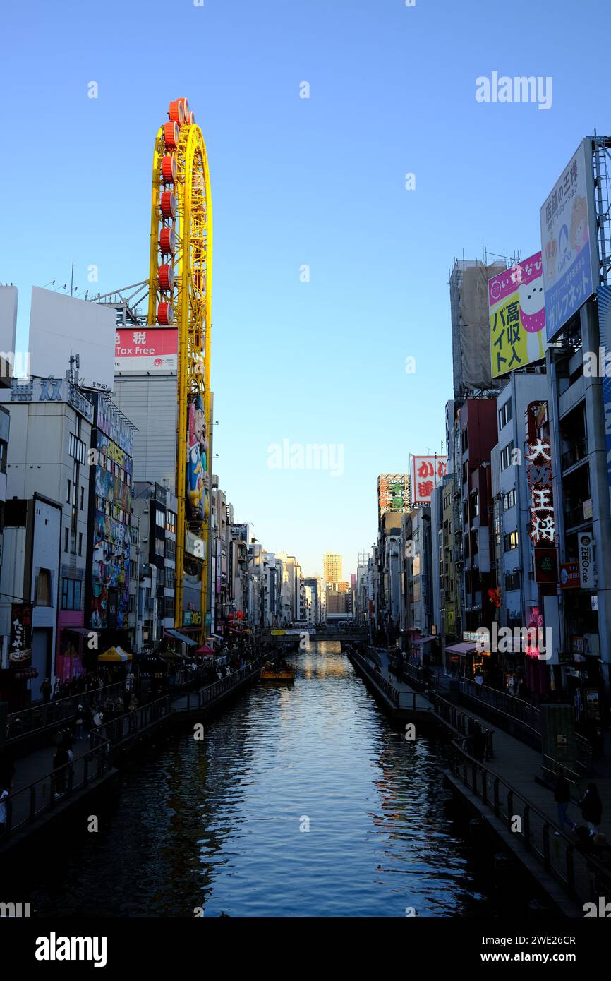 Lebendige urbane Kanalszene in Dotonbori, Osaka, mit farbenfrohen Anzeigen und Riesenrad vor klarem blauem Himmel. Stockfoto