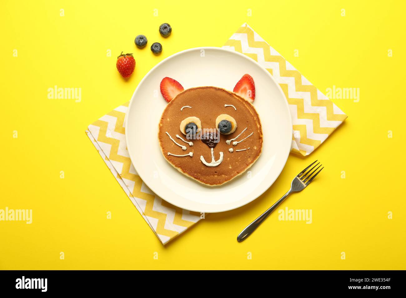 Kreatives Servieren für Kinder. Teller mit niedlicher Katze aus Pfannkuchen, Beeren, Creme, Banane und Schokoladenpaste auf gelbem Hintergrund, flach gelegt Stockfoto