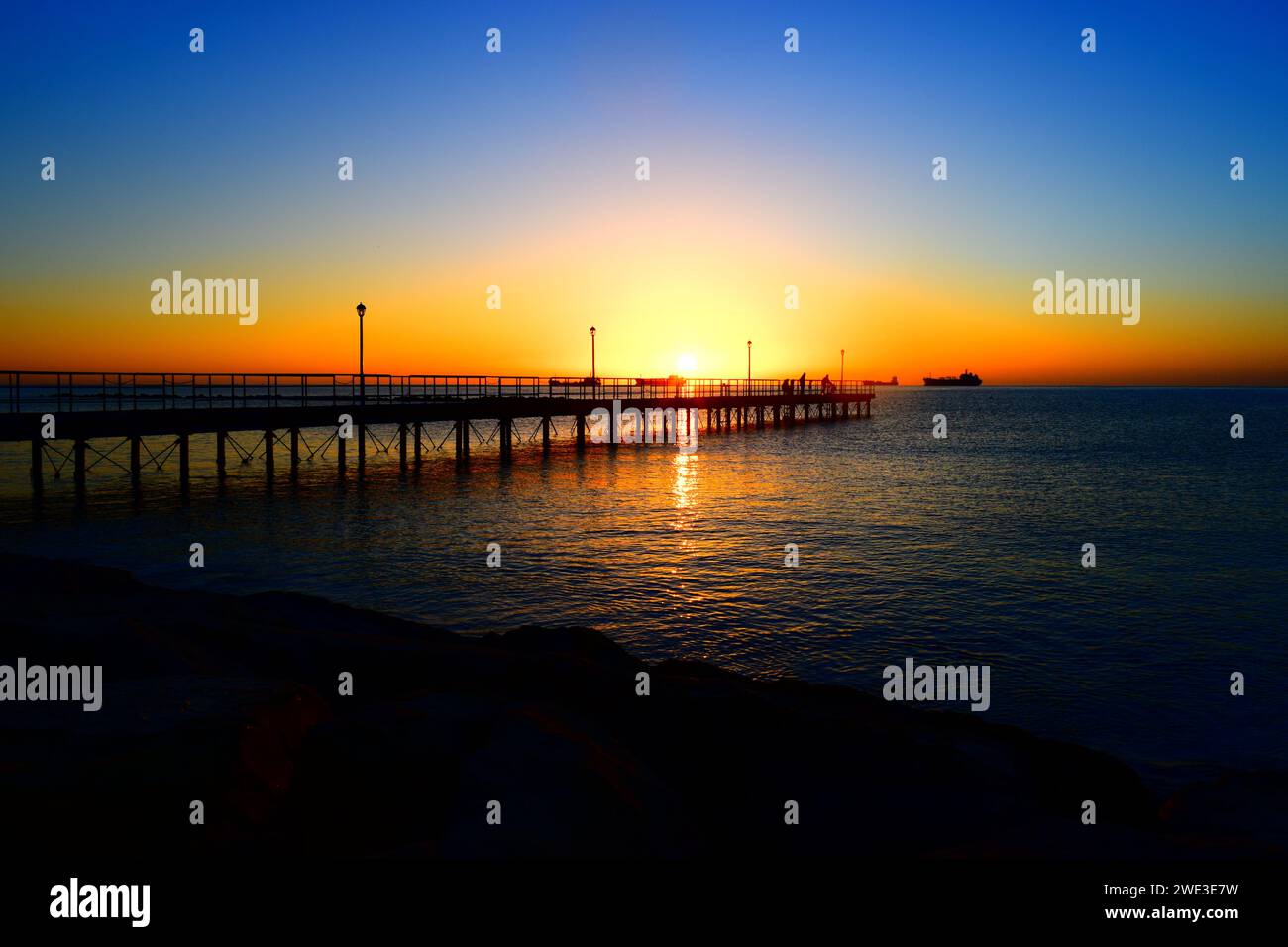 Ein atemberaubender Sonnenaufgang mit einem sehr großen Pier im Vordergrund und einem Schiff im Hintergrund. Stockfoto