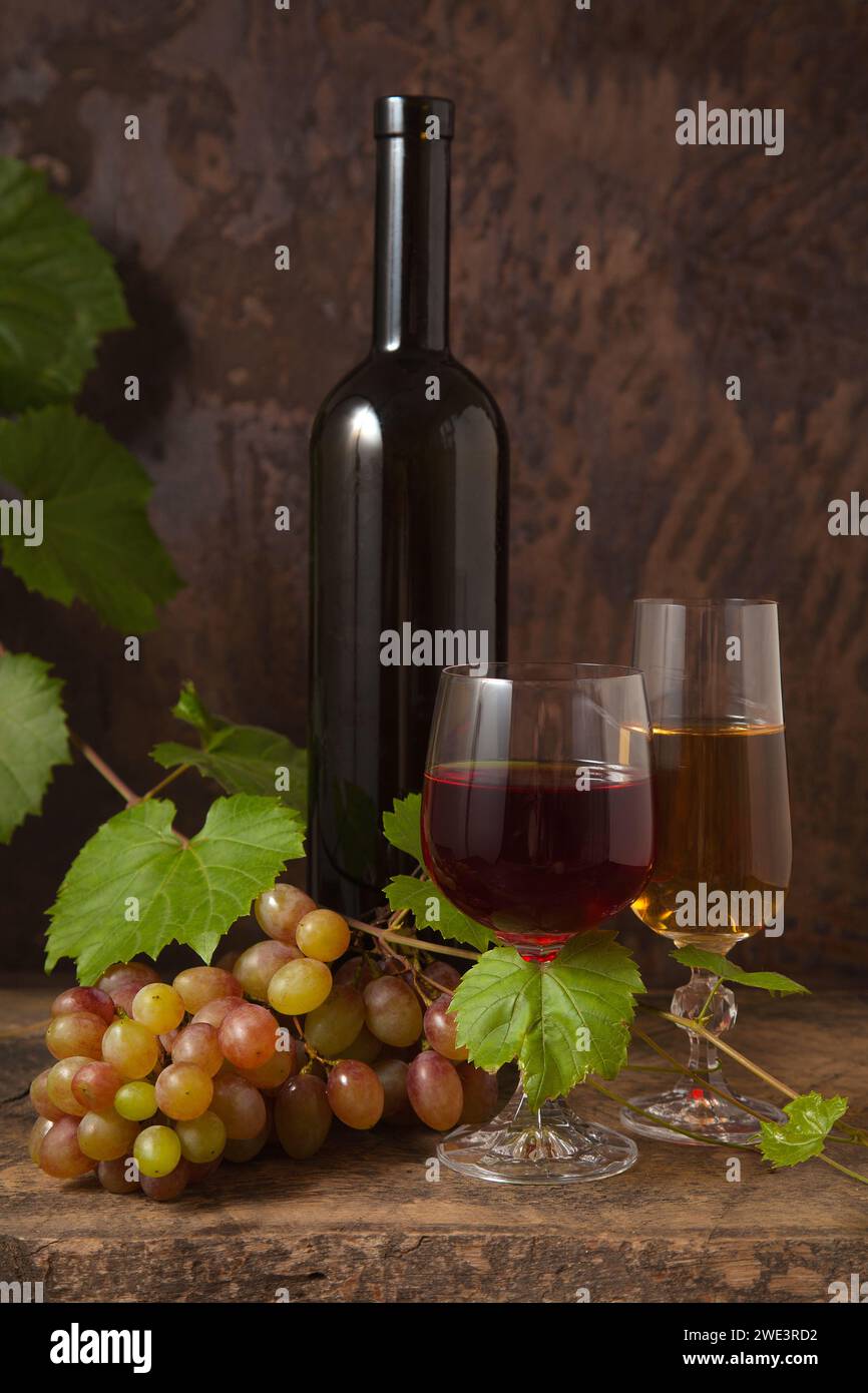 Das Konzept eines köstlichen alkoholischen Getränks, Wein und Trauben. Weinflasche, zwei Gläser Rot- und Weißwein und ein Haufen rosafarbener Trauben auf dunklem rustikalem Wo Stockfoto